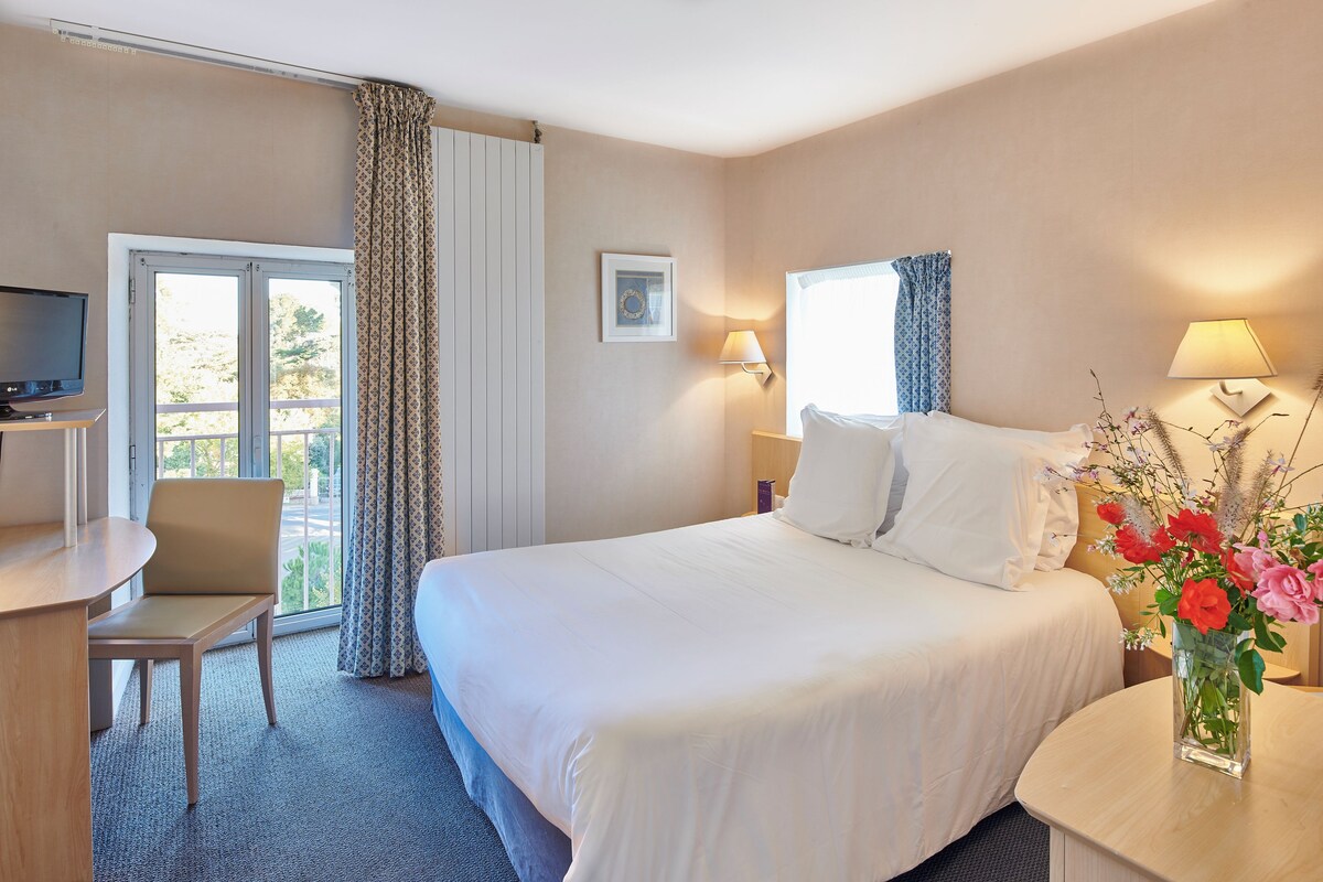 Ein Hotelzimmer mit Bett, Schreibtisch, Stuhl, Fernseher, einem Fenster mit Vorhängen, einer Wandlampe und einer Vase mit Blumen.