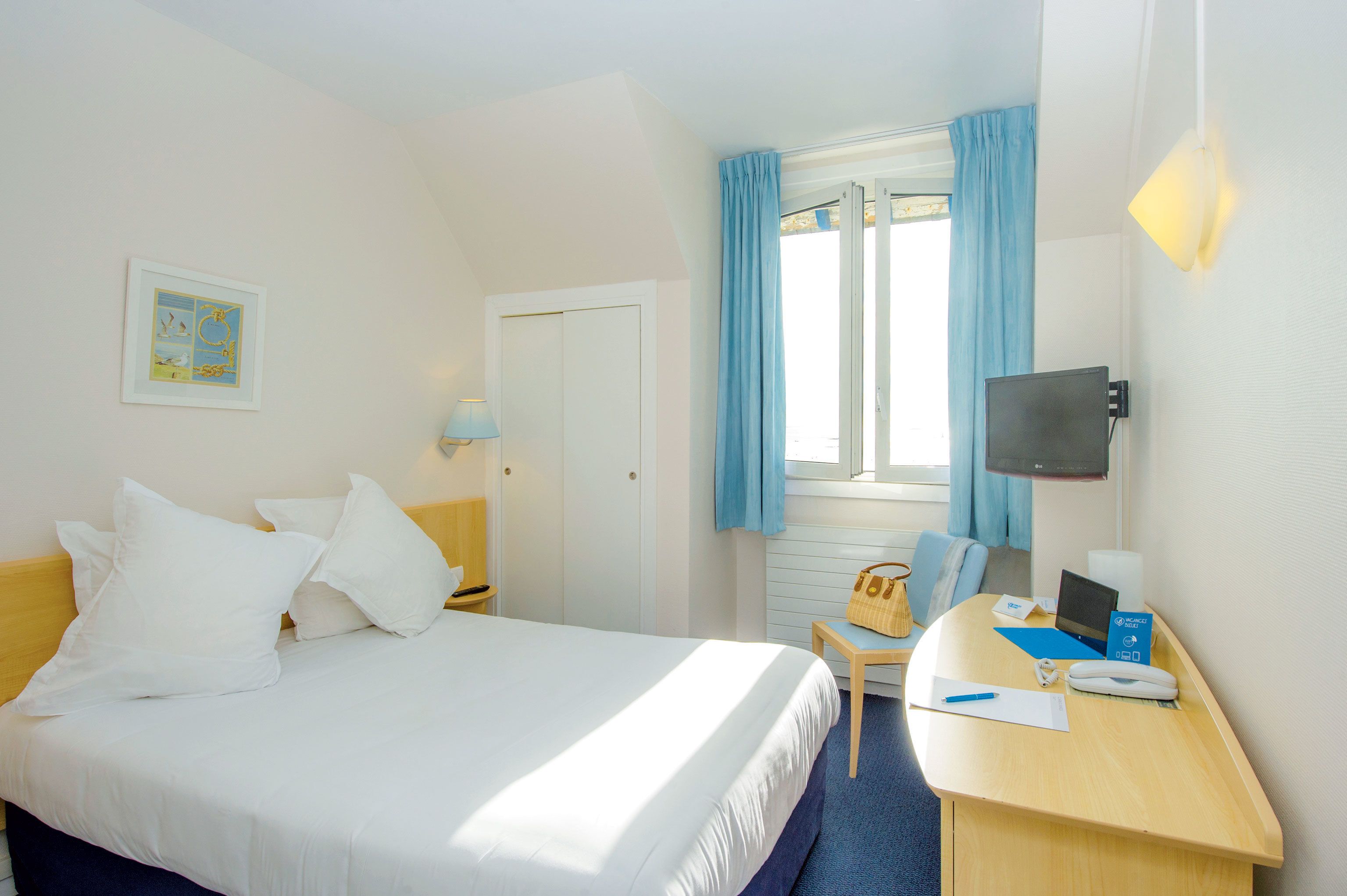 A hotel room with a bed, a desk with a laptop, a chair, a TV, and a window with curtains.