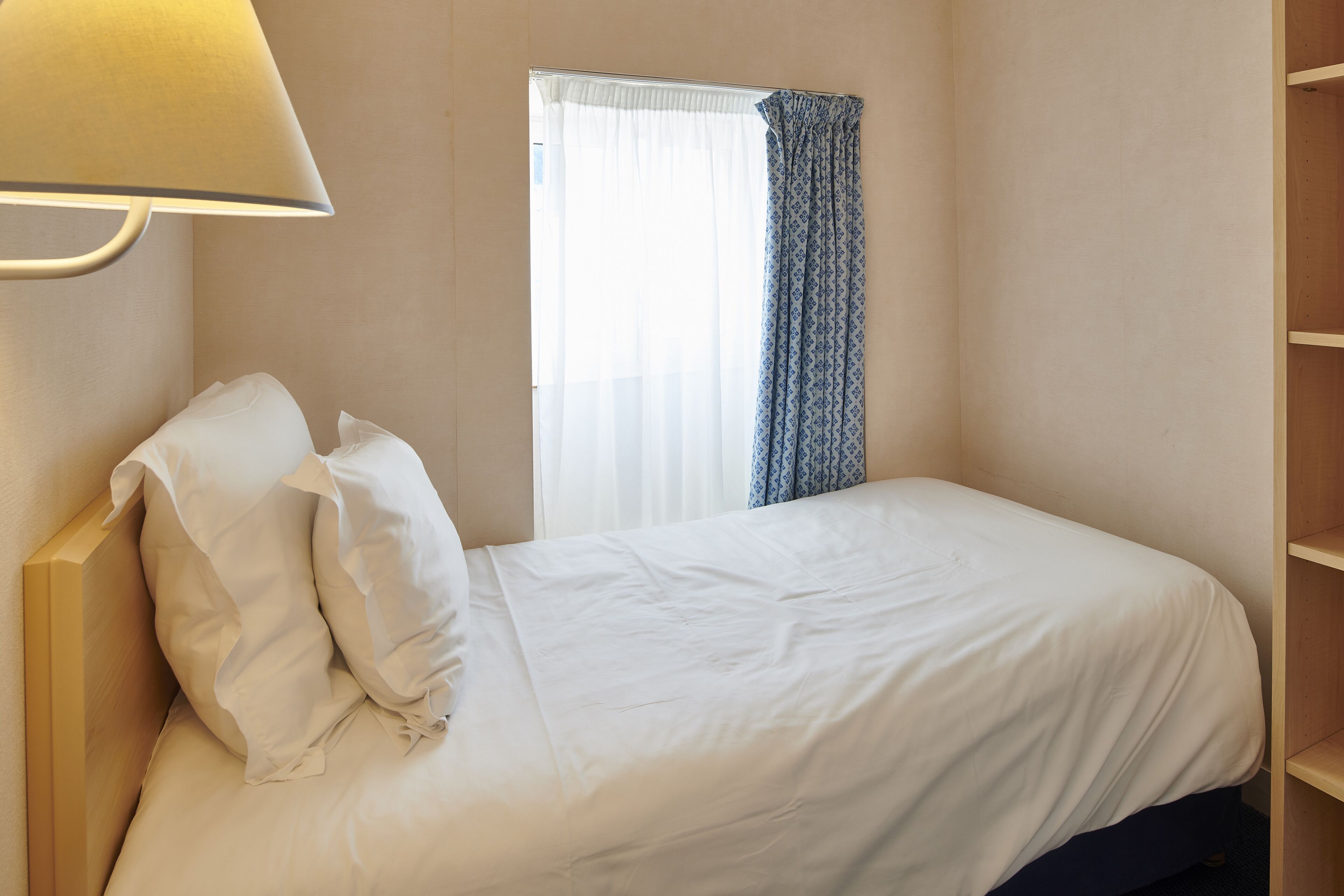 Un lit bien fait, avec du linge de lit blanc, une lampe de chevet et une fenêtre ornée de rideaux légers.