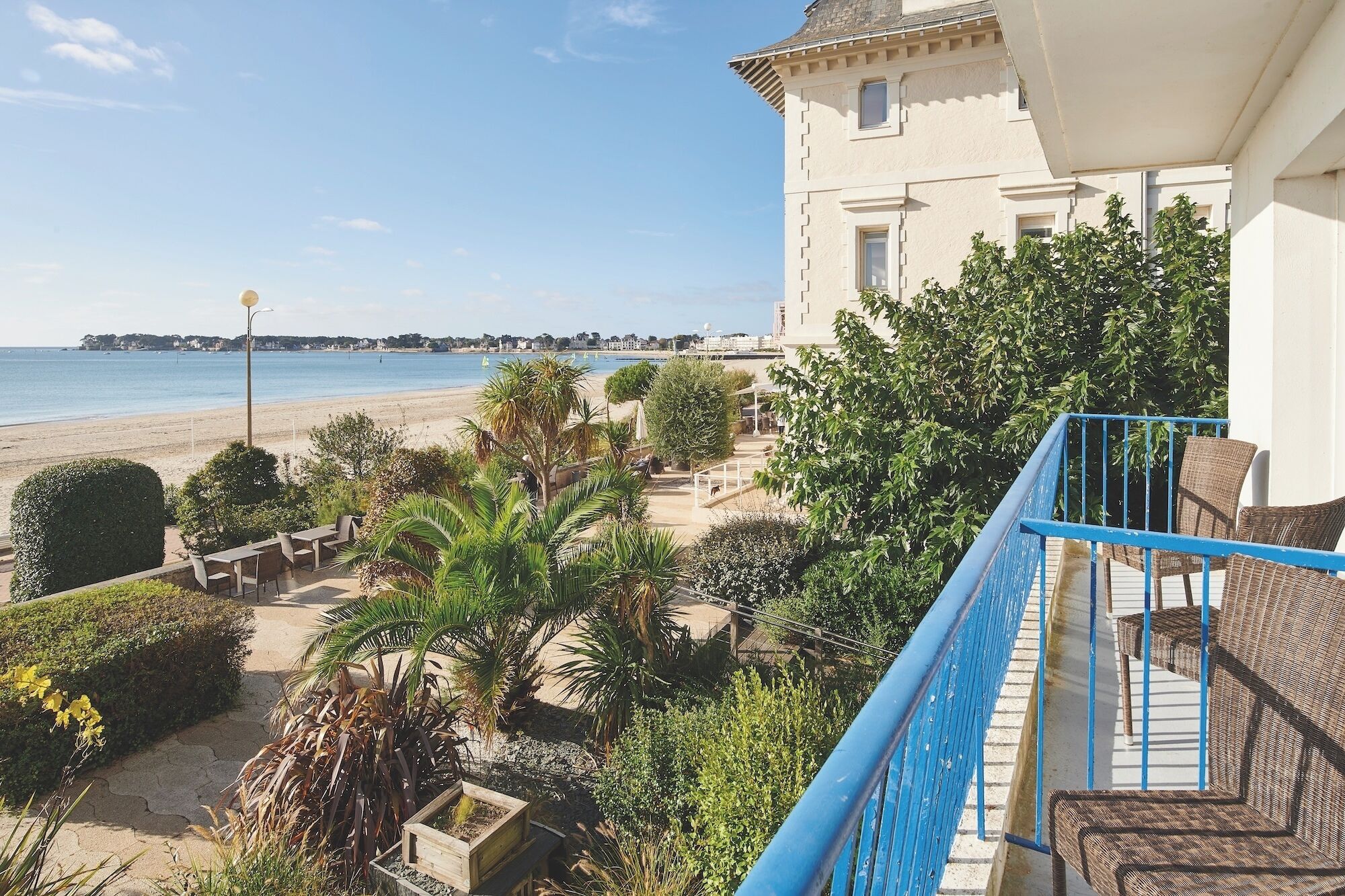 A coastal property with a balcony overlooking the beach, a garden with various plants, and a building with a white facade.