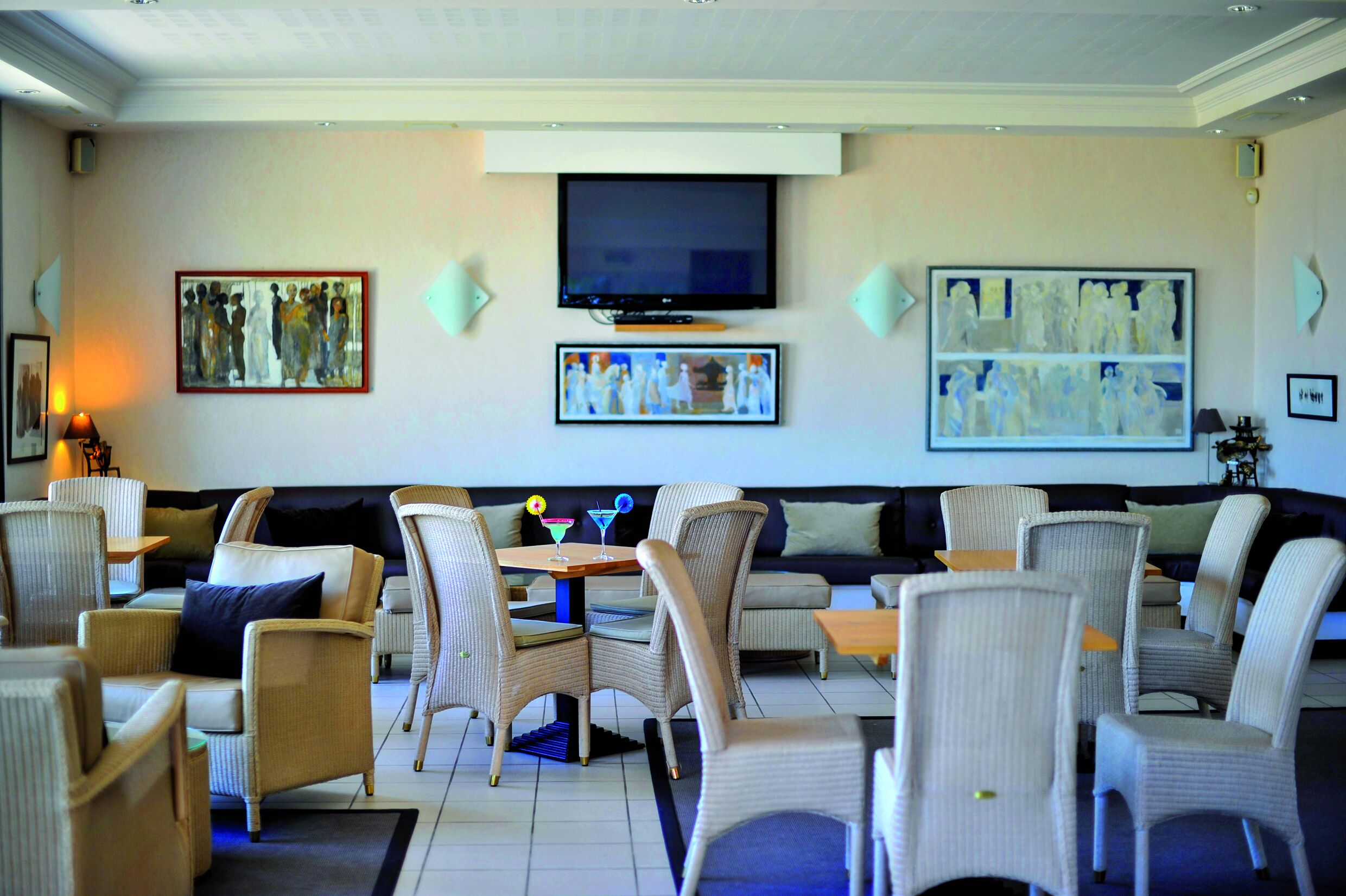 A hotel lobby with a seating area featuring wicker chairs and sofas, a flat-screen TV mounted on the wall, and artwork displayed on the walls.