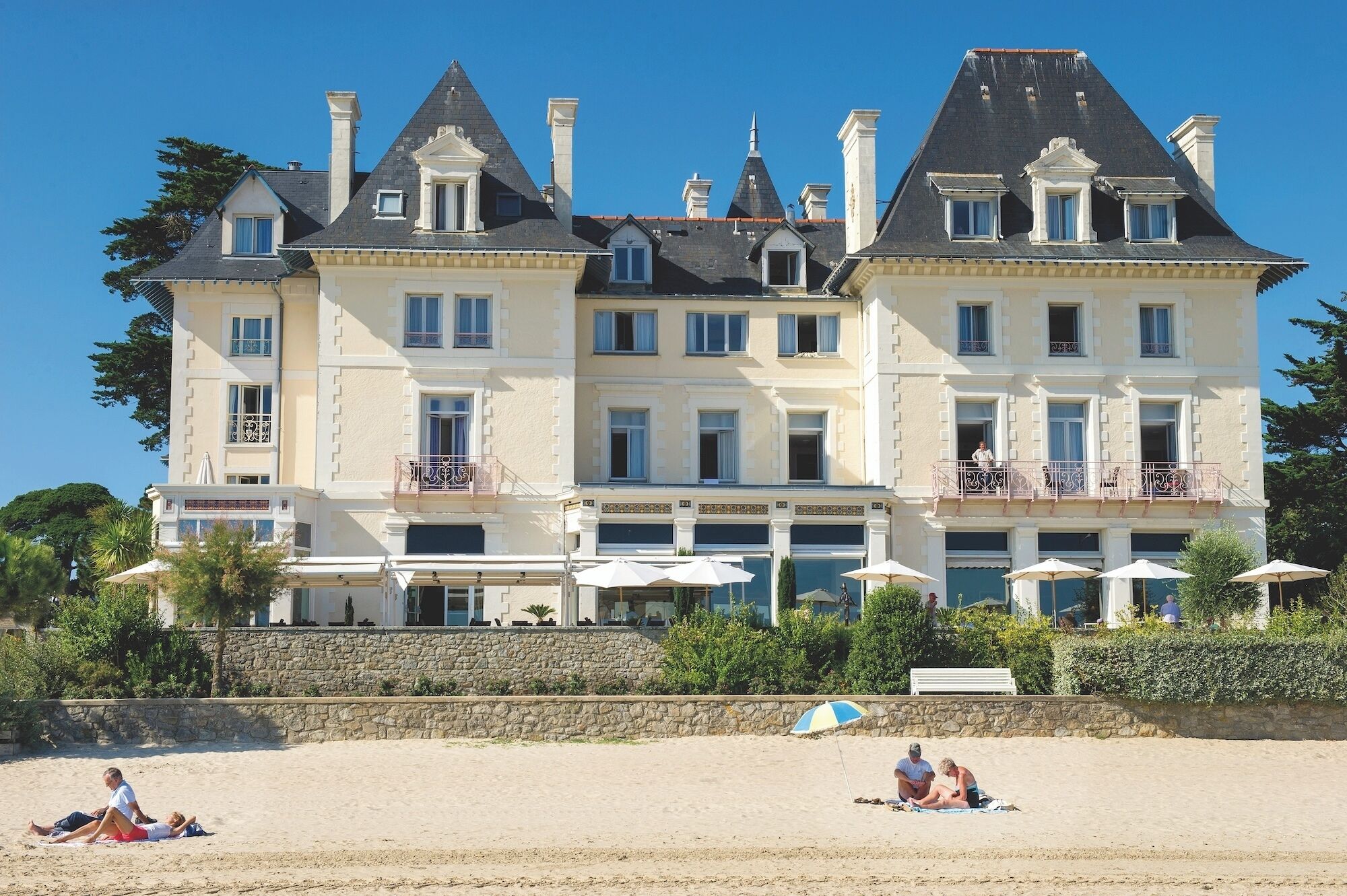 Ein großes, mehrstöckiges Gebäude mit einem markanten Balkonbereich, einer Steinwand im Vordergrund und einem Strand.
