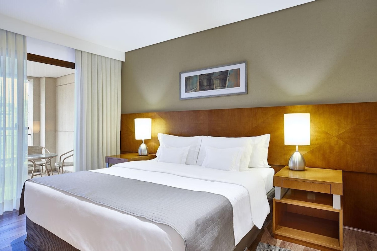 Una habitación de hotel con cama, dos lámparas de noche, un cuadro en la pared y vistas al exterior.
