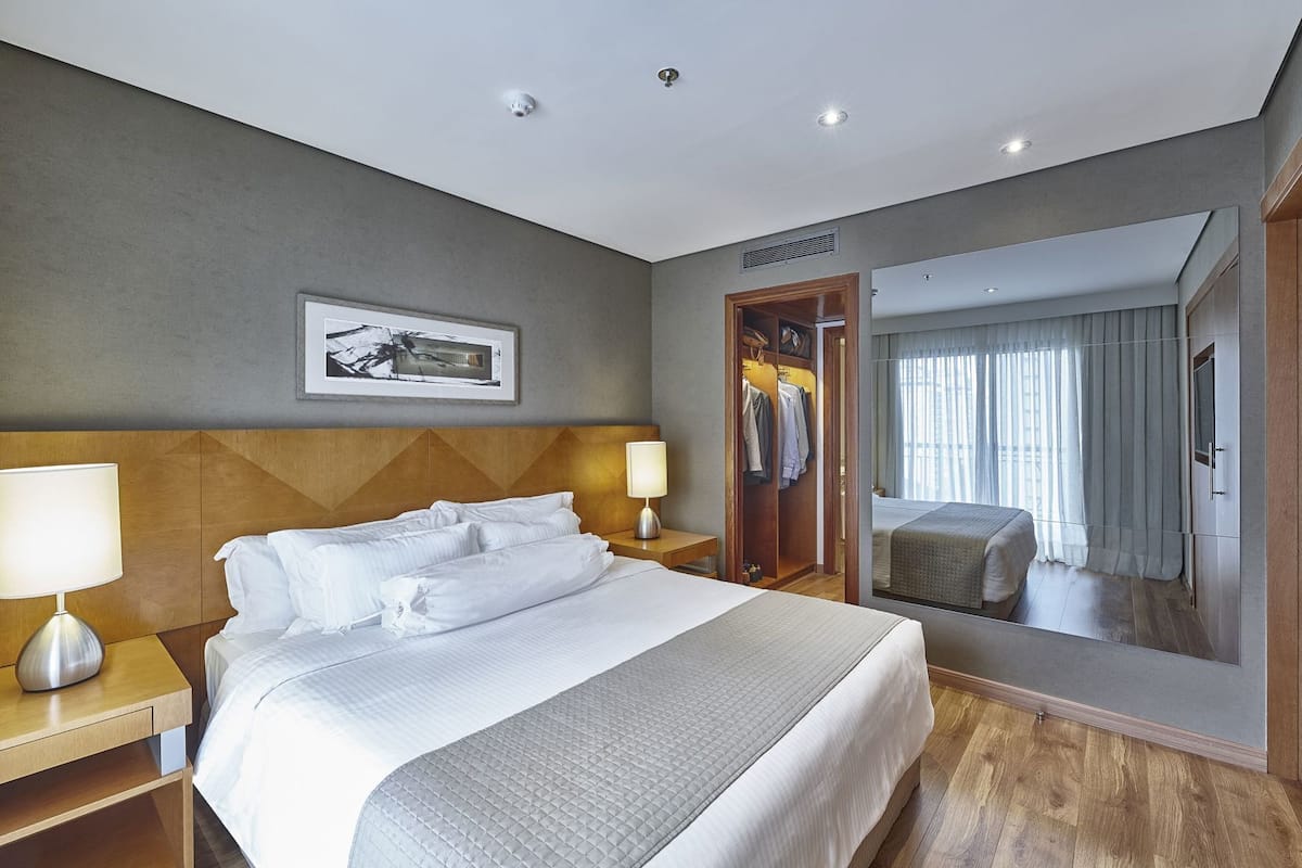 A hotel room with a large bed, two bedside tables, a wardrobe, a mirror, and a ceiling-mounted security camera.