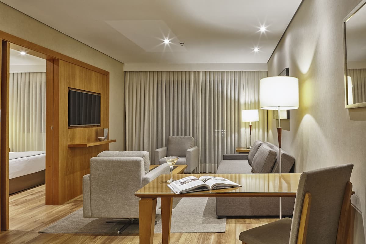 A hotel room with a wooden bed, a sofa, a dining table, and a TV mounted on the wall.