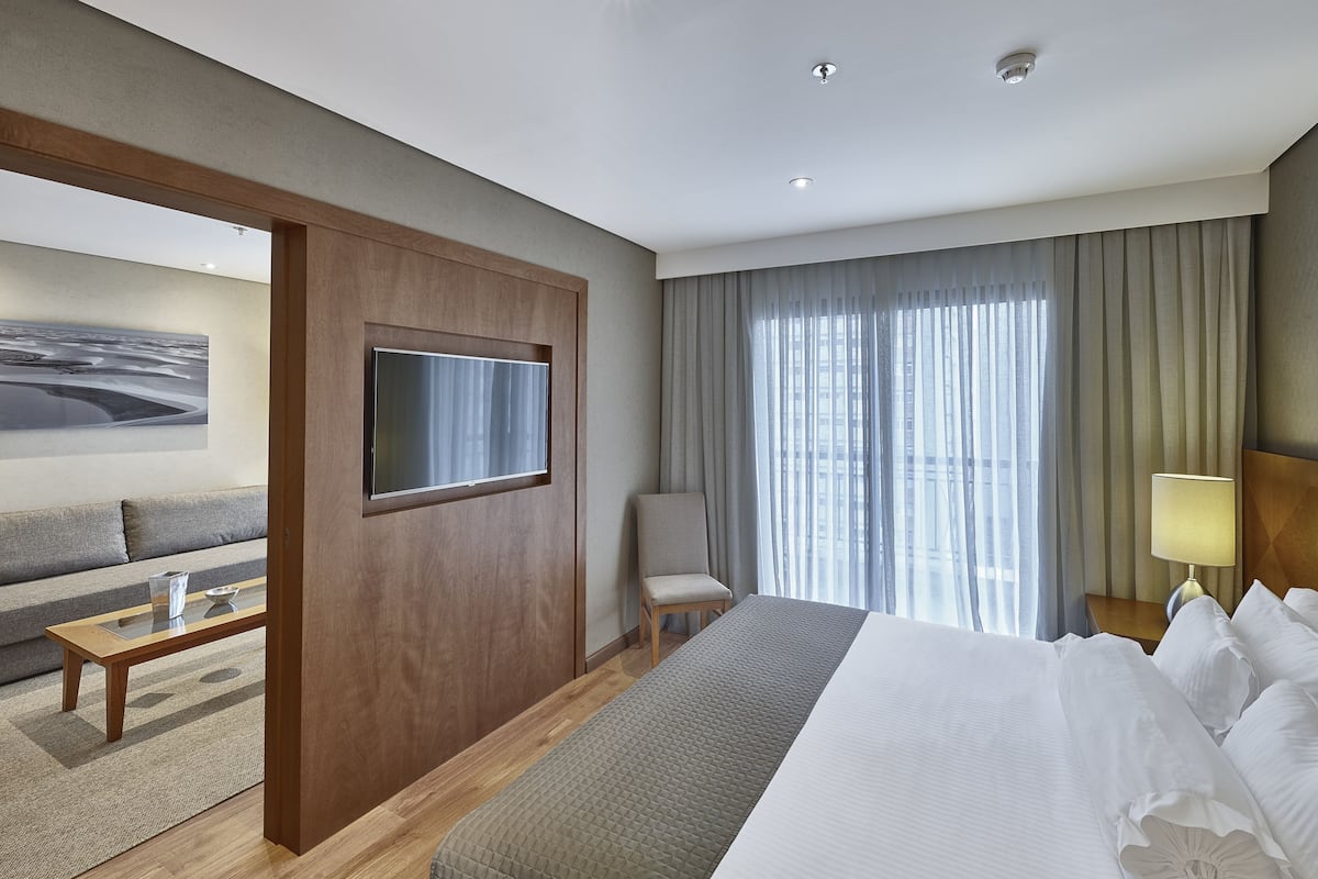 A hotel room with a bed, a TV mounted on the wall, a chair, a small table, and a window with curtains.