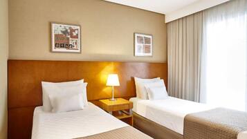 A hotel room with two beds, a headboard, a nightstand with a lamp, and a window with curtains.