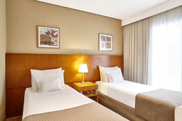 A hotel room with two beds, a headboard, a nightstand with a lamp, and a window with curtains. - George V Casa Branca (São Paulo)
