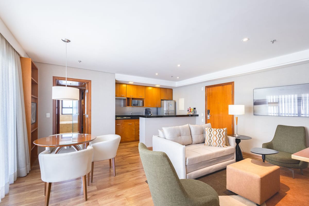 A modern hotel room with a living area, a dining table, a sofa, a green armchair, and a kitchen in the background.