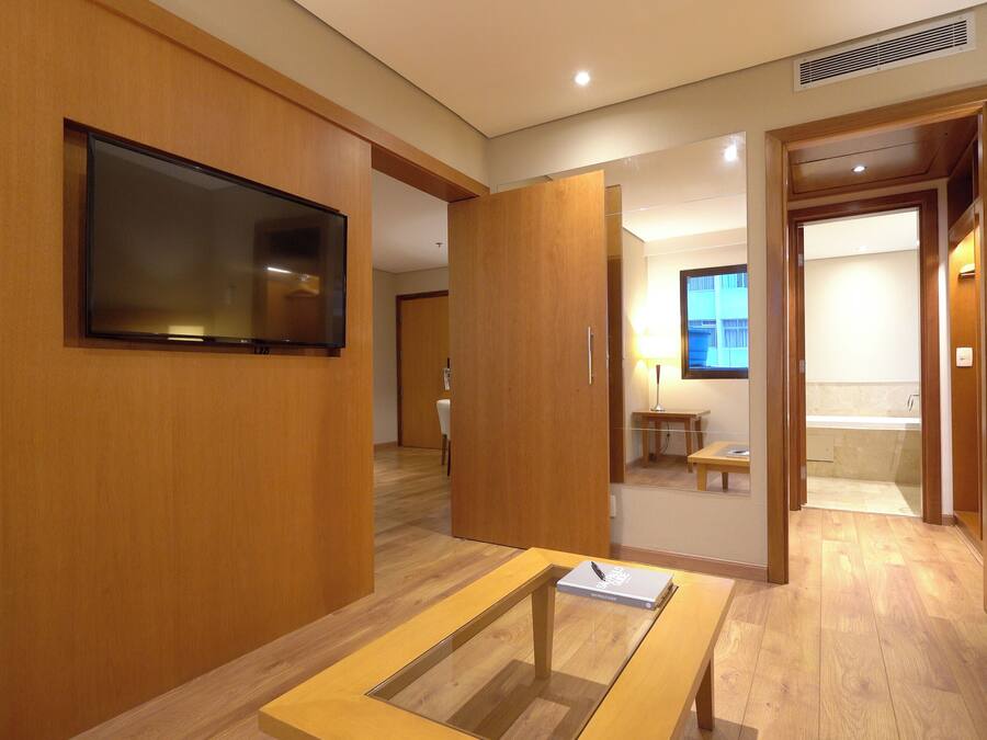 A hotel room with a wooden floor, a flat-screen TV mounted on the wall, a small table with a book, and a sofa.