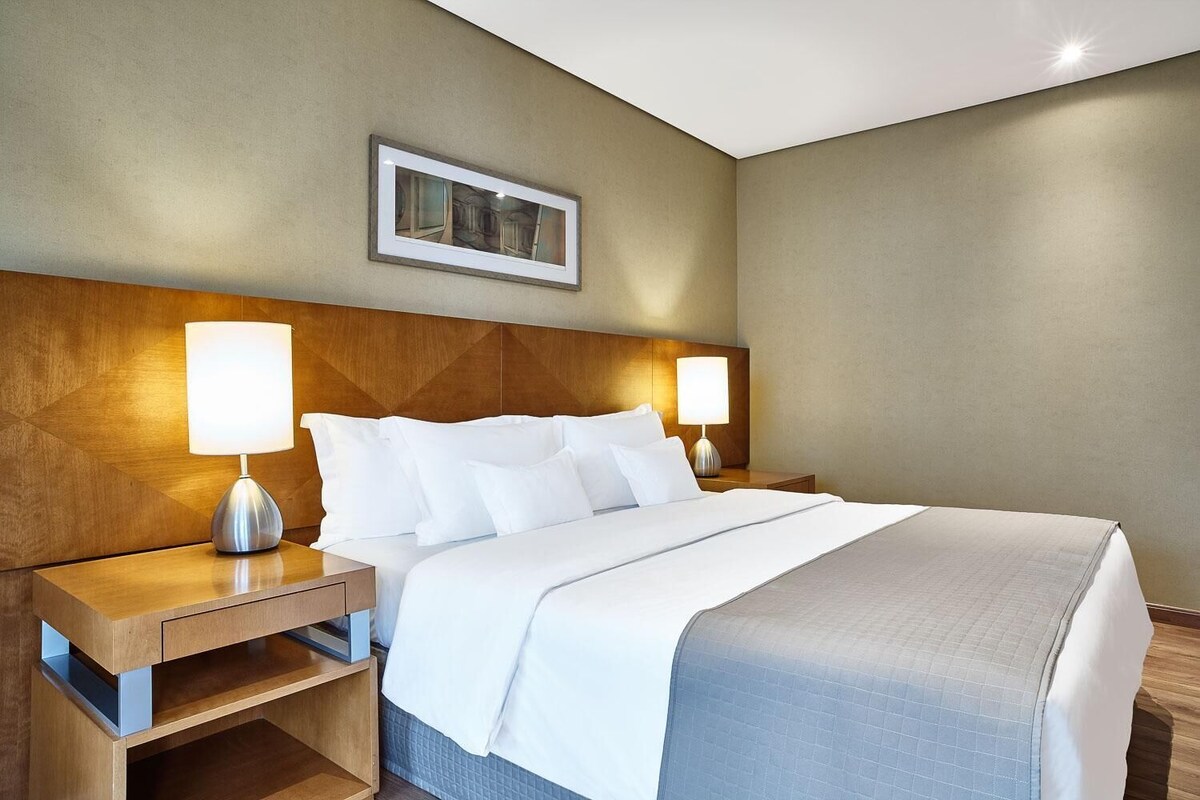 Habitación de hotel con una cama, dos mesitas de noche con lámparas, un cabecero de madera, un cuadro enmarcado en la pared y una colcha azul acolchada.