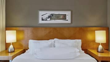 Quarto de hotel com cabeceira de madeira, duas mesas de cabeceira com luminárias, cama com roupa de cama branca e um quadro emoldurado na parede.