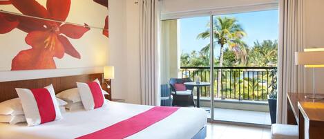 A hotel room with a large bed, a balcony with a view of palm trees, and a dining area with a table and chairs.