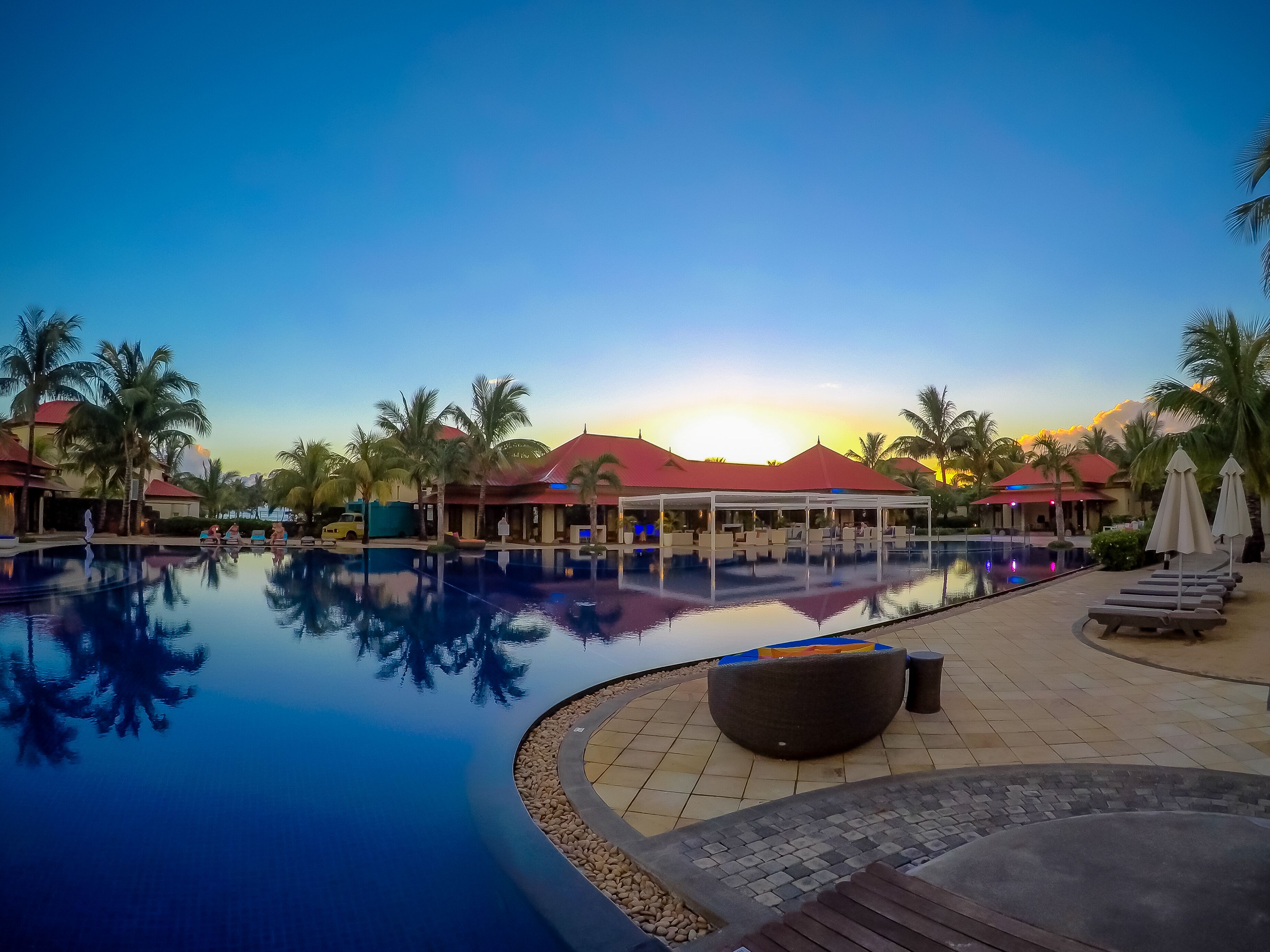 A resort pool with a clear view of the sunset, palm trees, and a covered seating area.