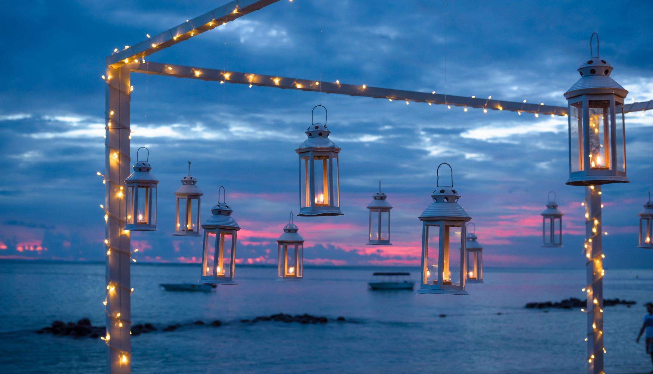 A beach scene with hanging lanterns and string lights against a twilight sky.