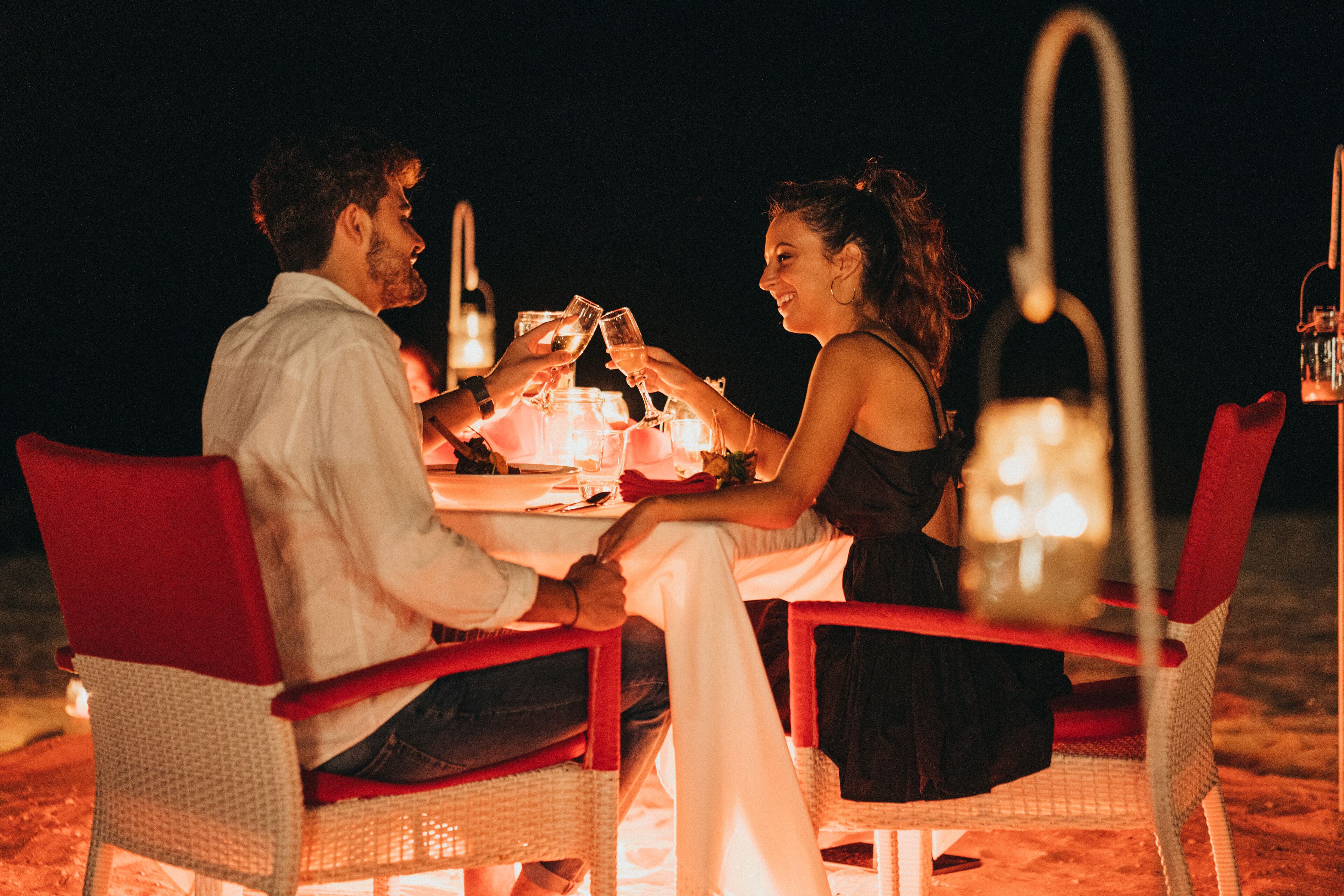 A couple dining at a table with red chairs, surrounded by candles and bottles, at night.