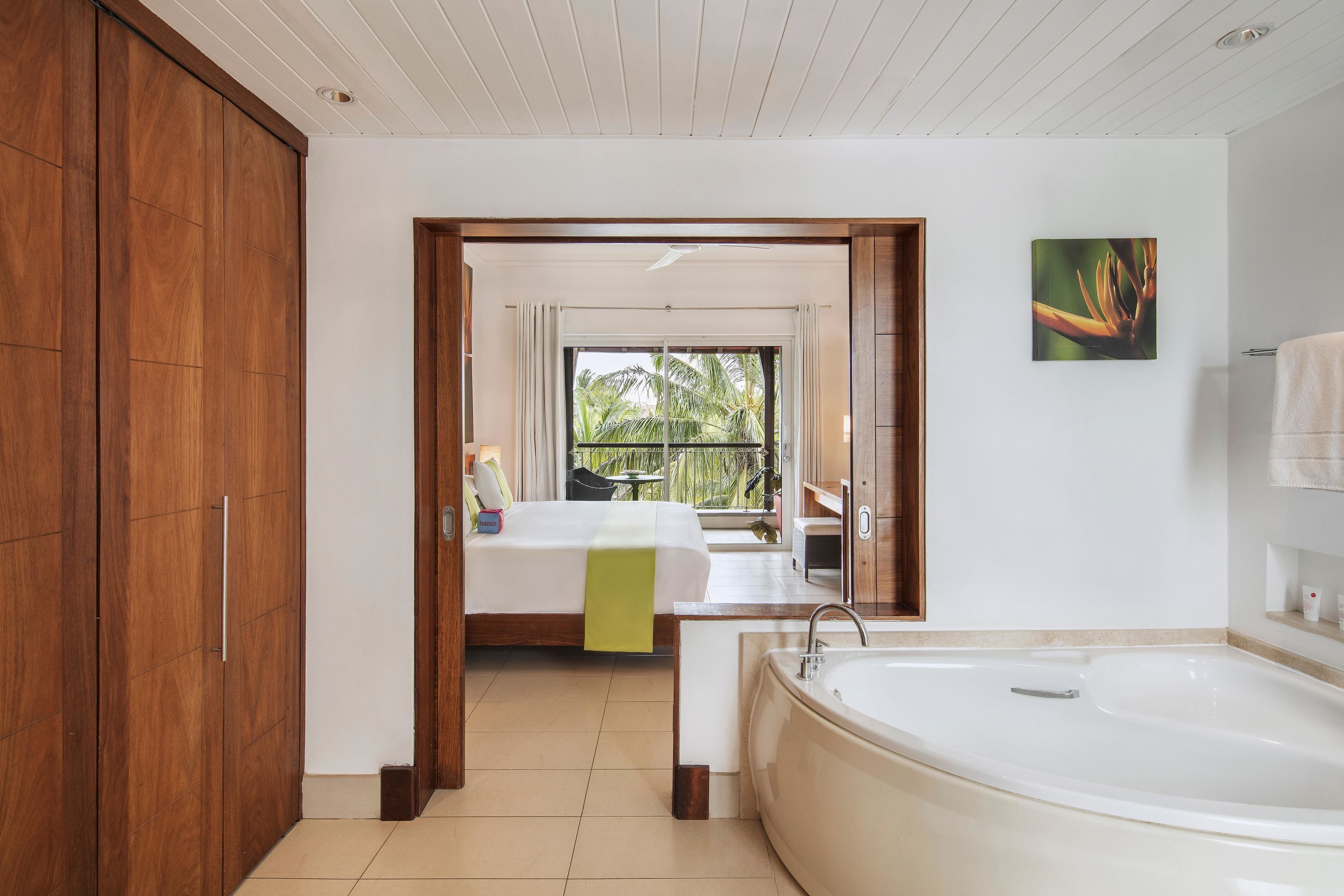 Una habitación de hotel moderna con una bañera grande, una cama con un plaid verde y un balcón con vistas a palmeras.