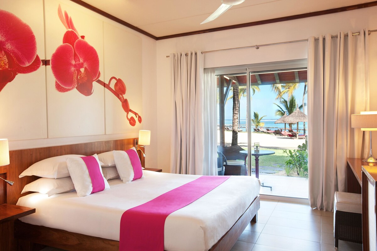 A hotel room with a bed, a desk, a chair, a window with a view of the beach, and a tropical-themed wall decoration.