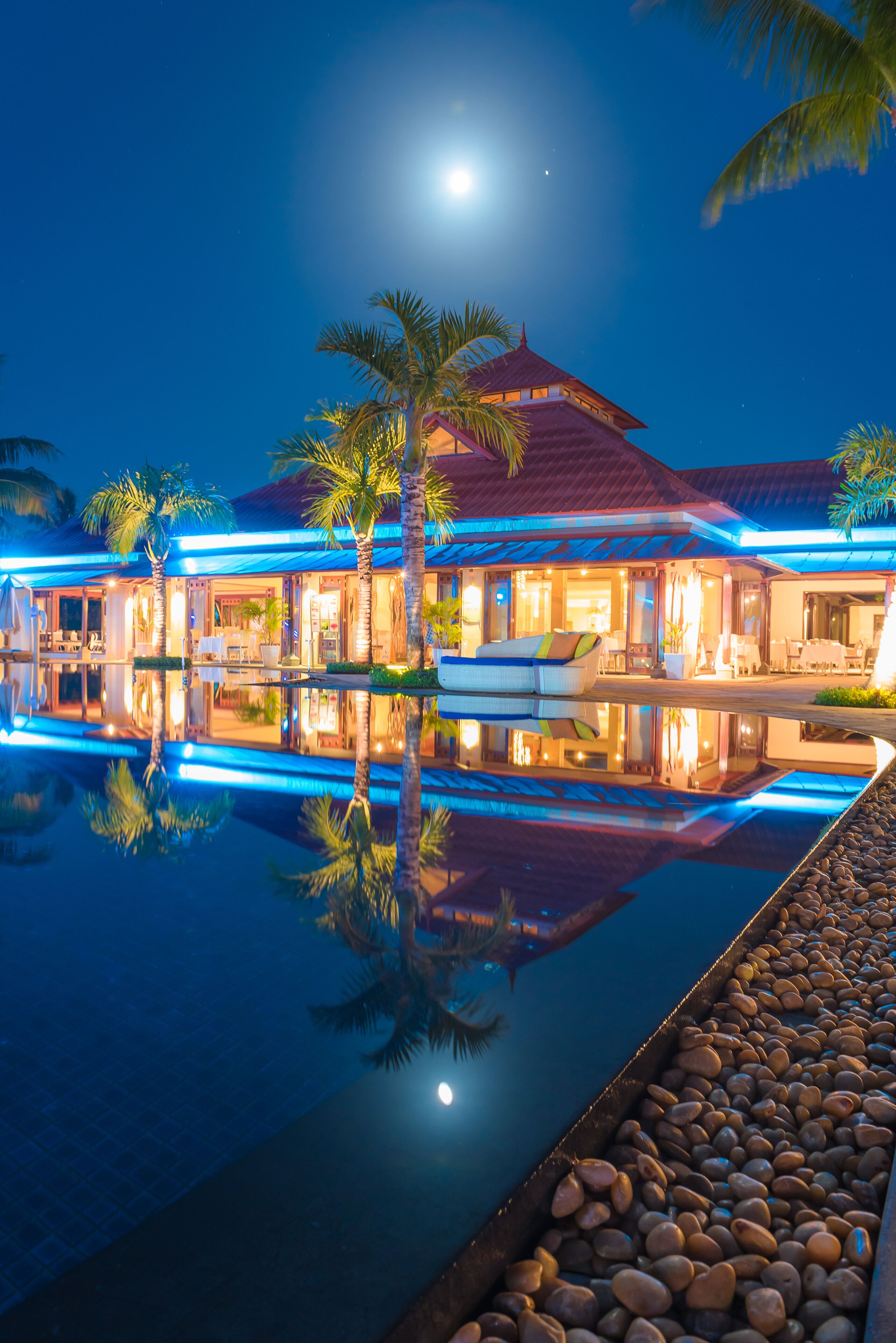 A resort with a swimming pool reflecting the building and palm trees, a covered outdoor seating area, and a well-lit entrance.