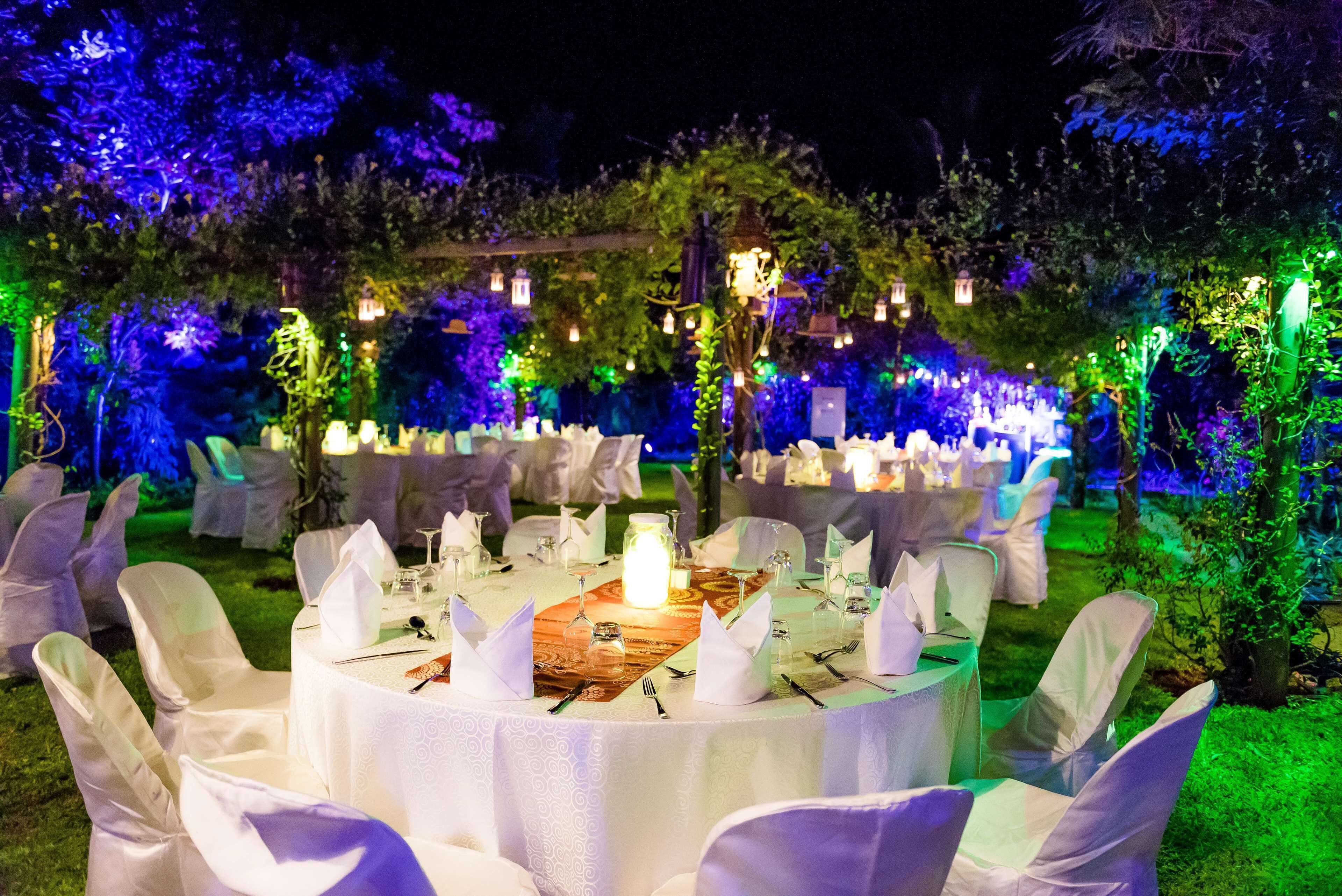 Mesa redonda dispuesta para un evento al aire libre formal, con manteles blancos y sillas, rodeada de follaje e iluminación decorativa.