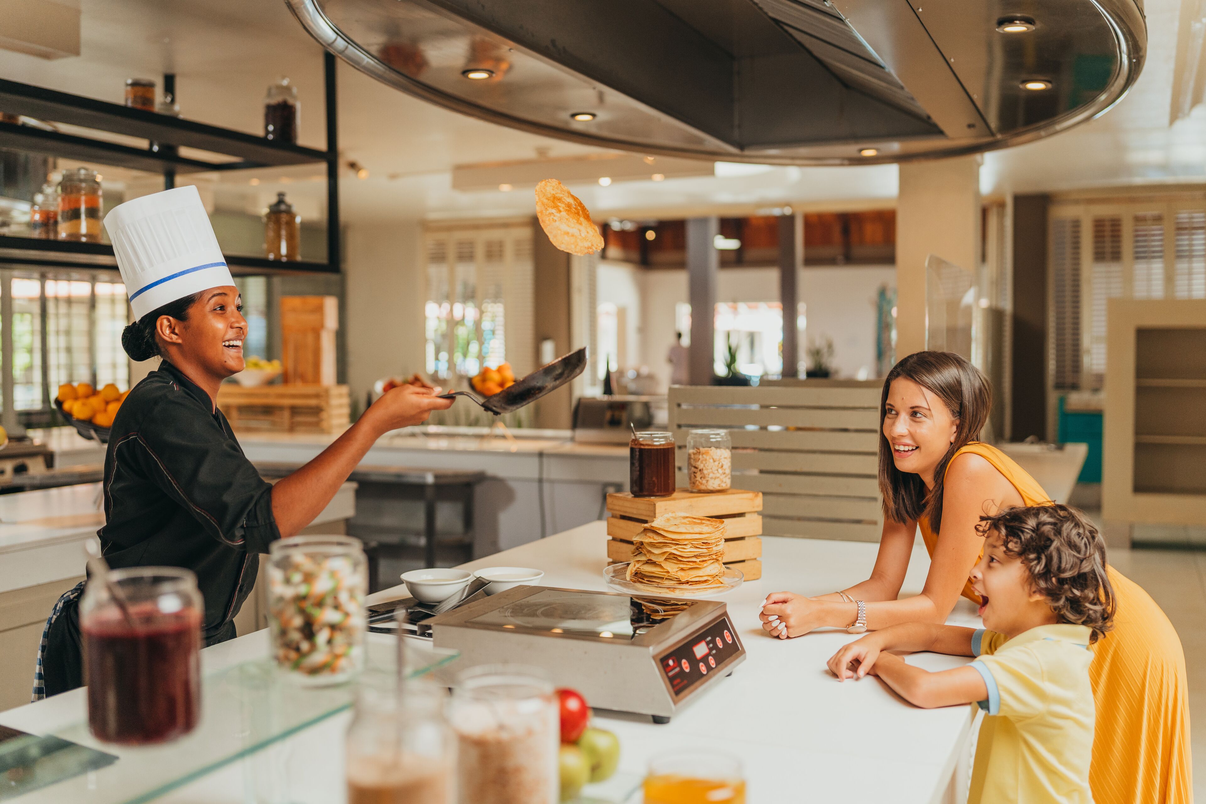 Une cuisine moderne où un chef prépare des plats, une table à manger pour la famille, ainsi que divers appareils de cuisine et ustensiles.