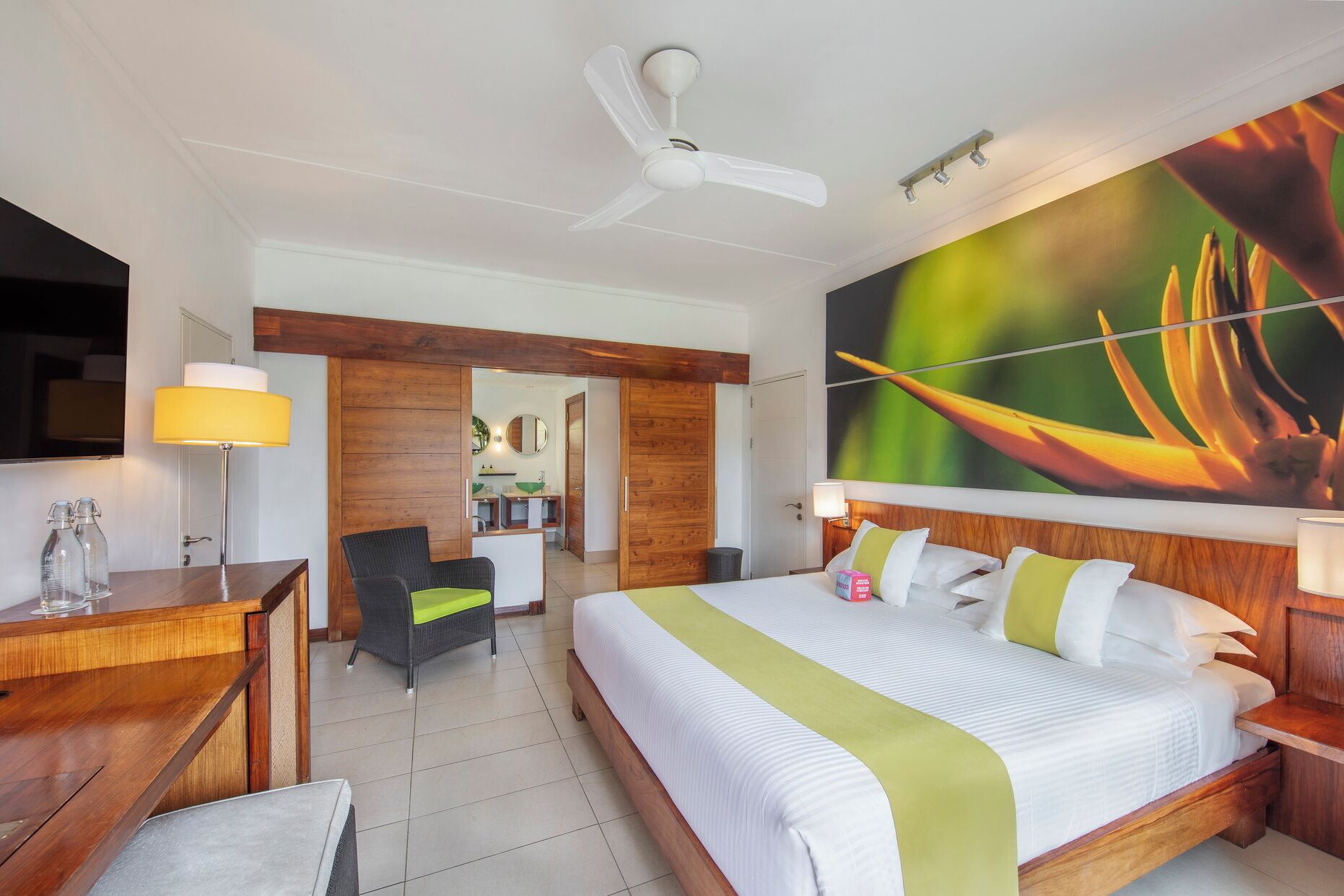 A hotel room with a bed, a desk, a chair, a TV, a ceiling fan, and a bathroom visible through an open door.