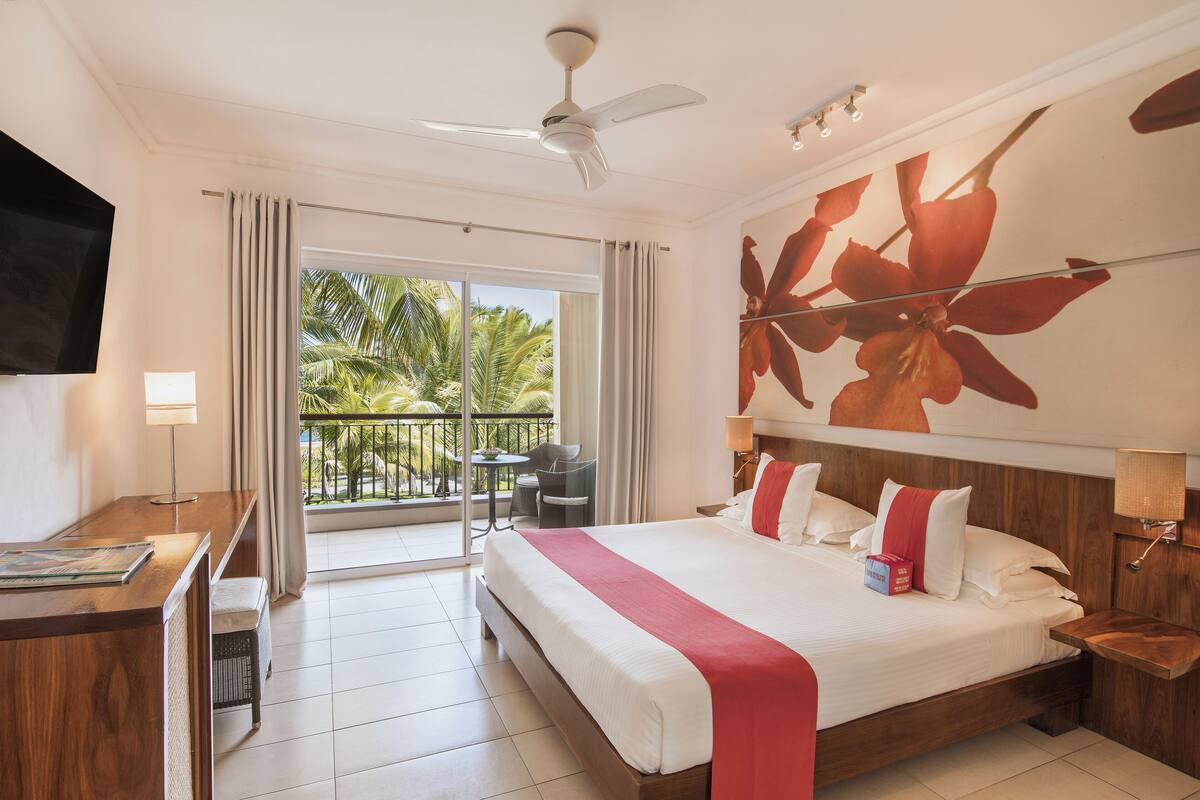 Une chambre d’hôtel avec un grand lit, un bureau, une chaise, une télévision et un balcon offrant une vue sur les palmiers.