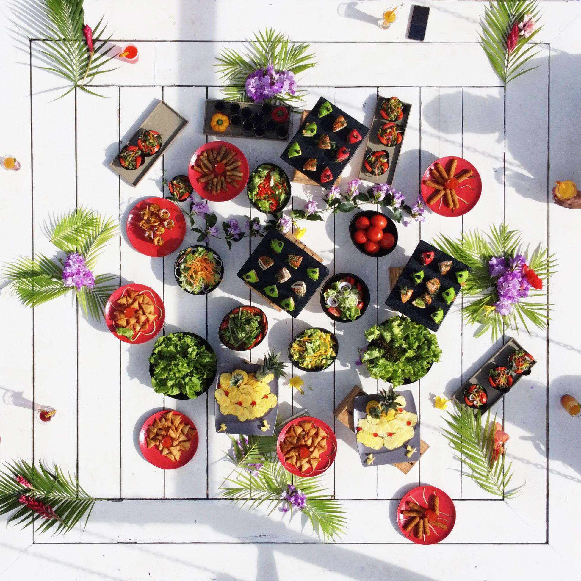 Vue d’ensemble d’un espace de restauration, avec différentes assiettes et boissons disposées sur une table en bois blanche, le tout agrémenté de plantes et de fleurs décoratives.