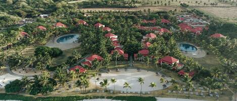 An aerial view of a resort with multiple red-roofed buildings, palm trees, and two swimming pools.