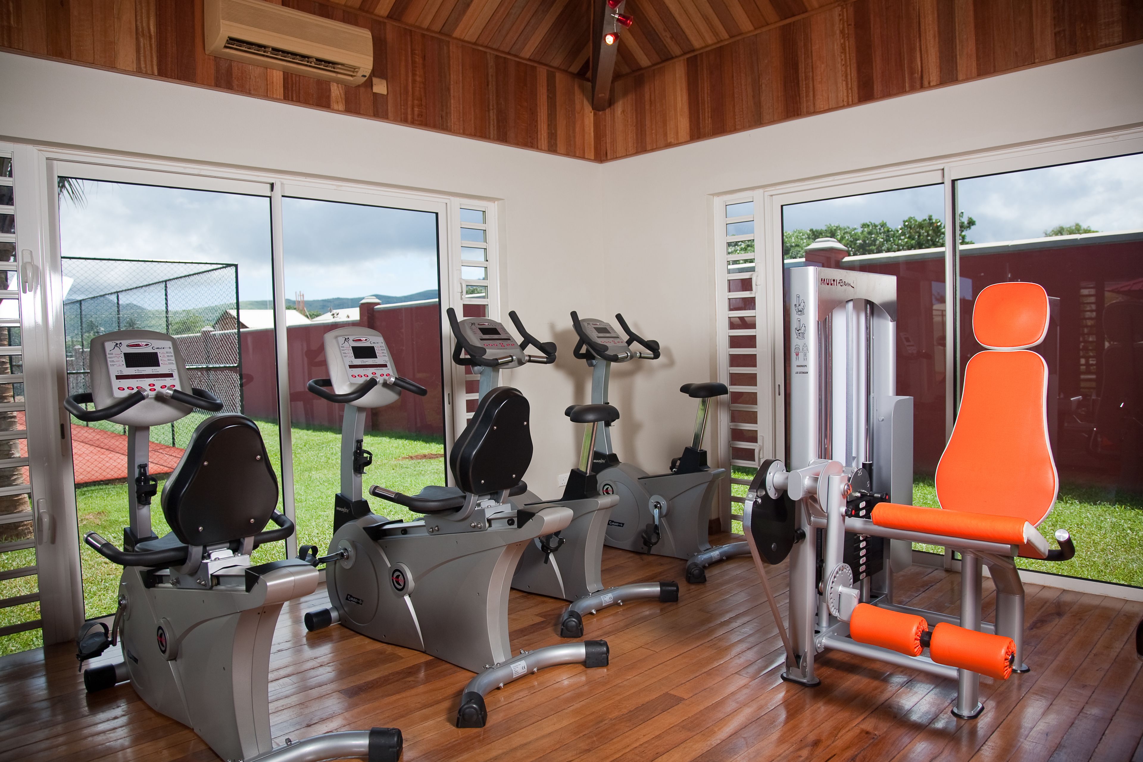 A gym with various exercise machines, large windows, and a wooden floor.