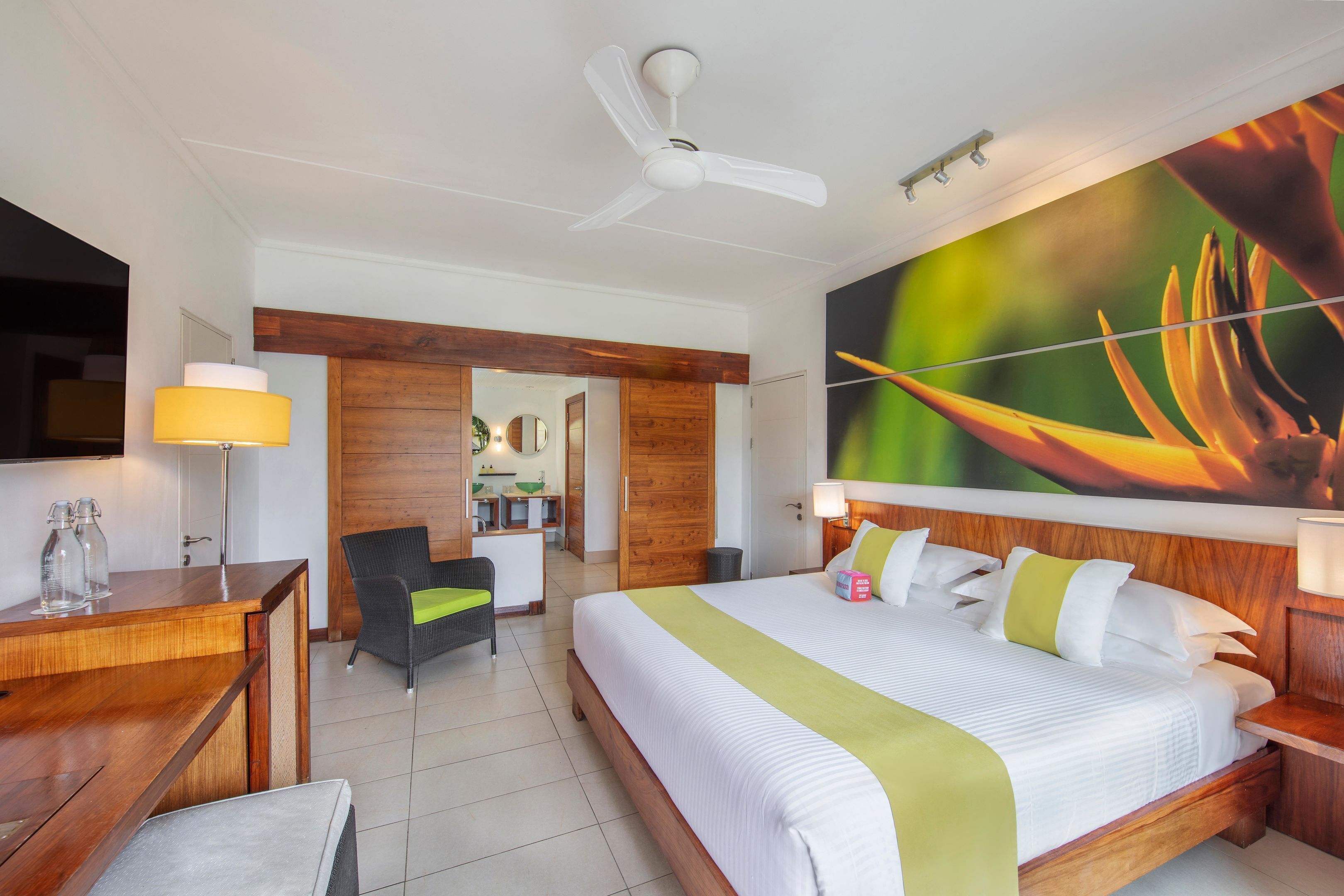 A hotel room with a bed, a desk, a chair, a ceiling fan, and a large wall painting.