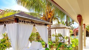 Un complejo turístico con zonas de estar al aire libre cubiertas por toldos blancos, rodeado de exuberante vegetación tropical y una piscina.