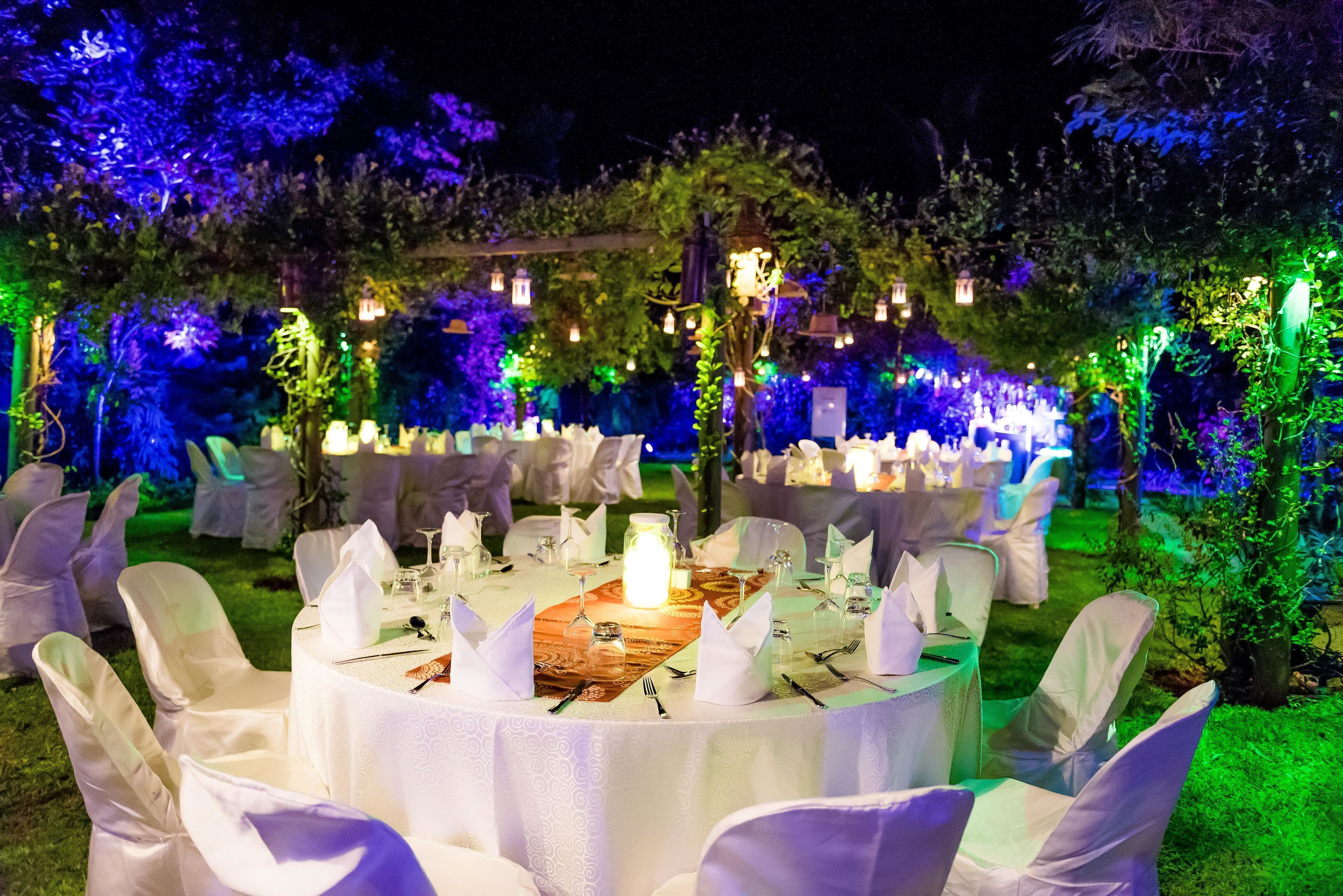 An outdoor dining area with round tables, white tablecloths, and chairs, surrounded by greenery and string lights.