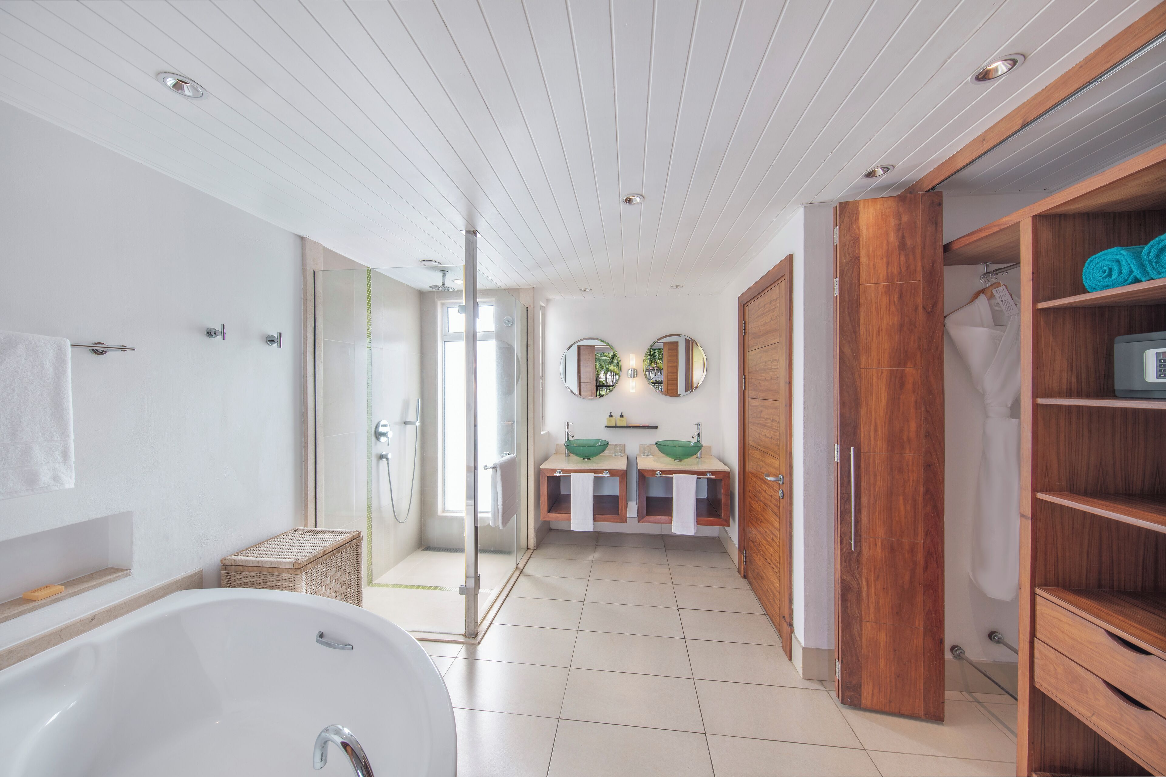 A modern bathroom with a freestanding bathtub, two sinks with green bowls, a walk-in shower, and a wooden vanity with shelves.