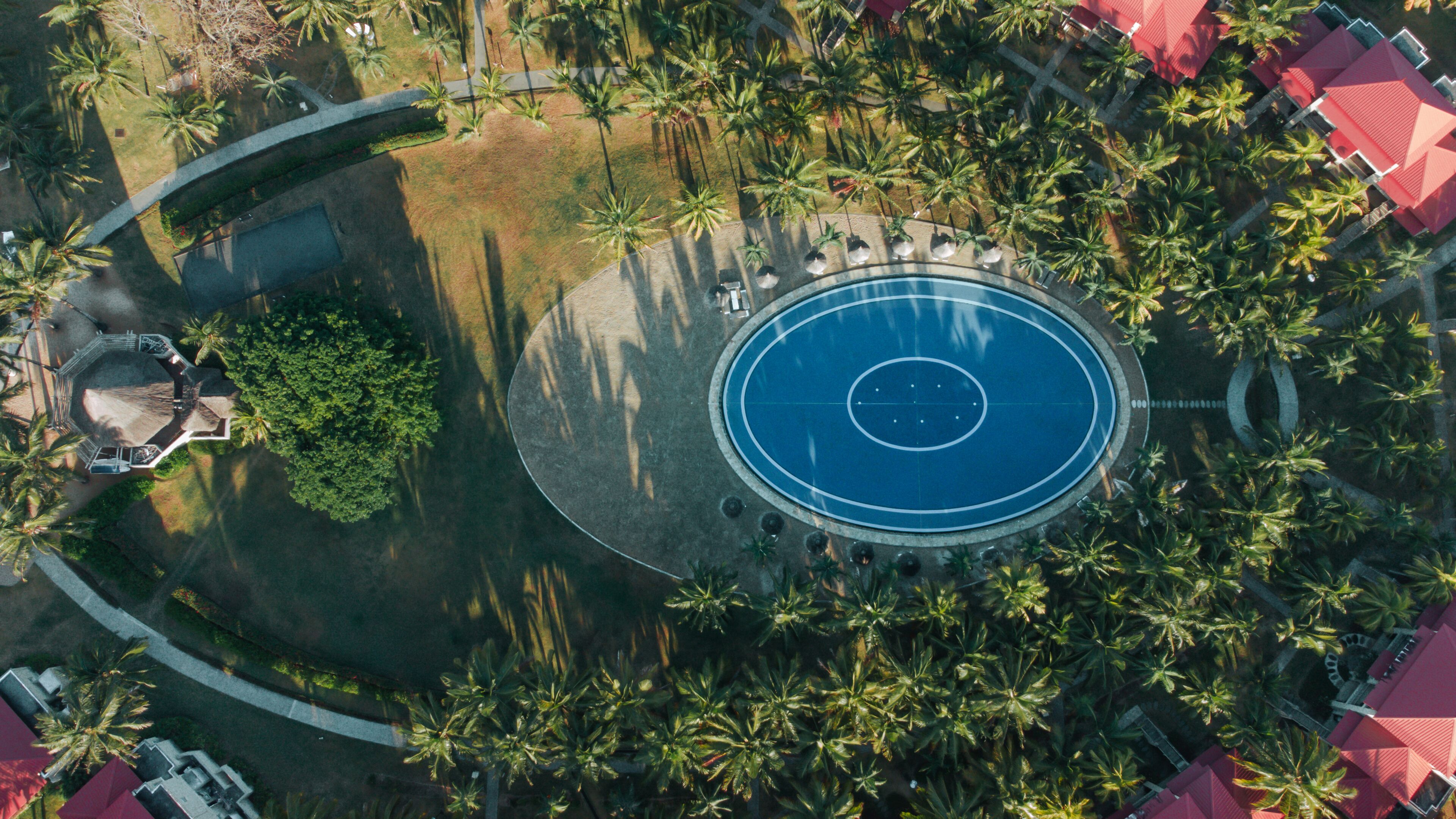 An aerial view of a resort with a central circular pool, surrounded by palm trees and red-roofed structures.
