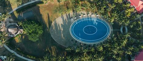 An aerial view of a resort with a central circular pool, surrounded by palm trees and red-roofed structures.