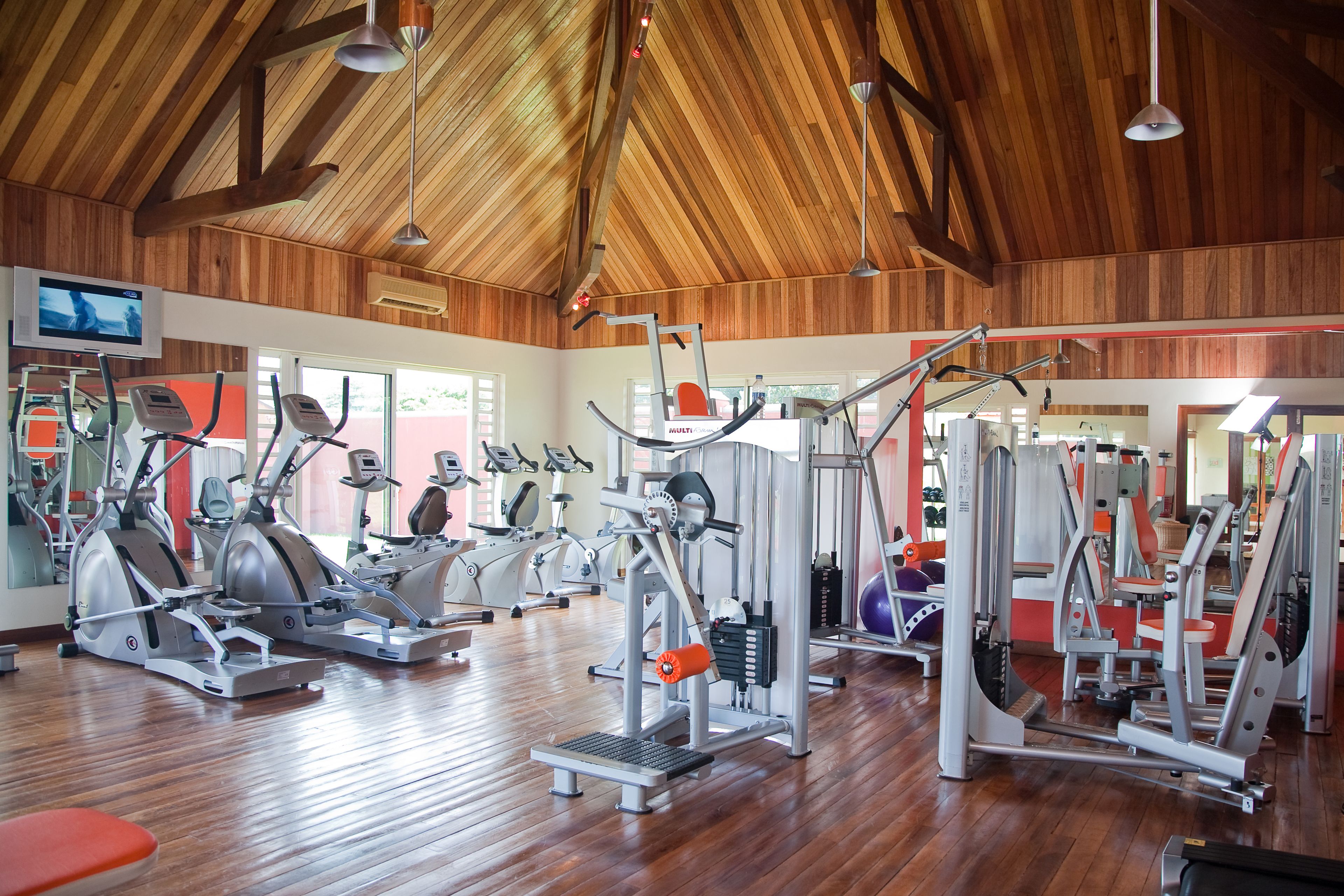 Un gimnasio con diverso equipo de ejercicio, vigas de techo de madera y un televisor en la pared.