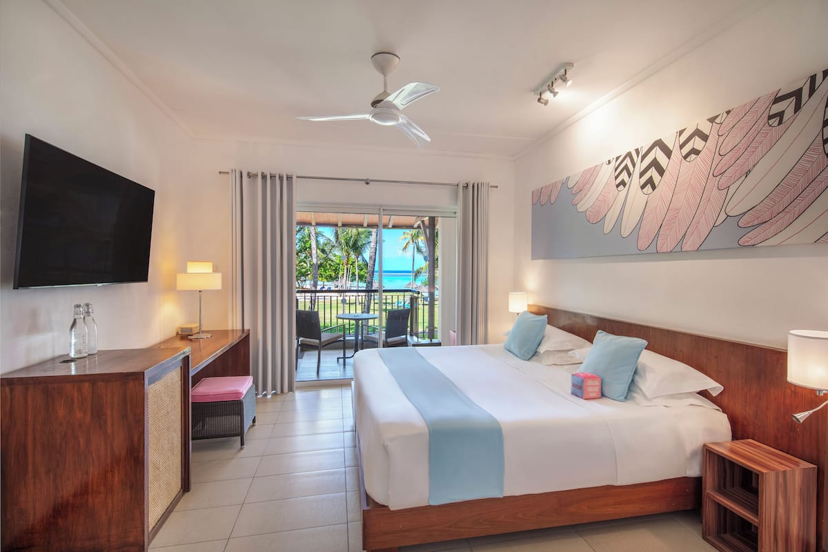 Une chambre d’hôtel équipée d’un lit, d’un bureau, d’une télévision, d’un balcon avec vue sur l’océan et d’un ventilateur de plafond.