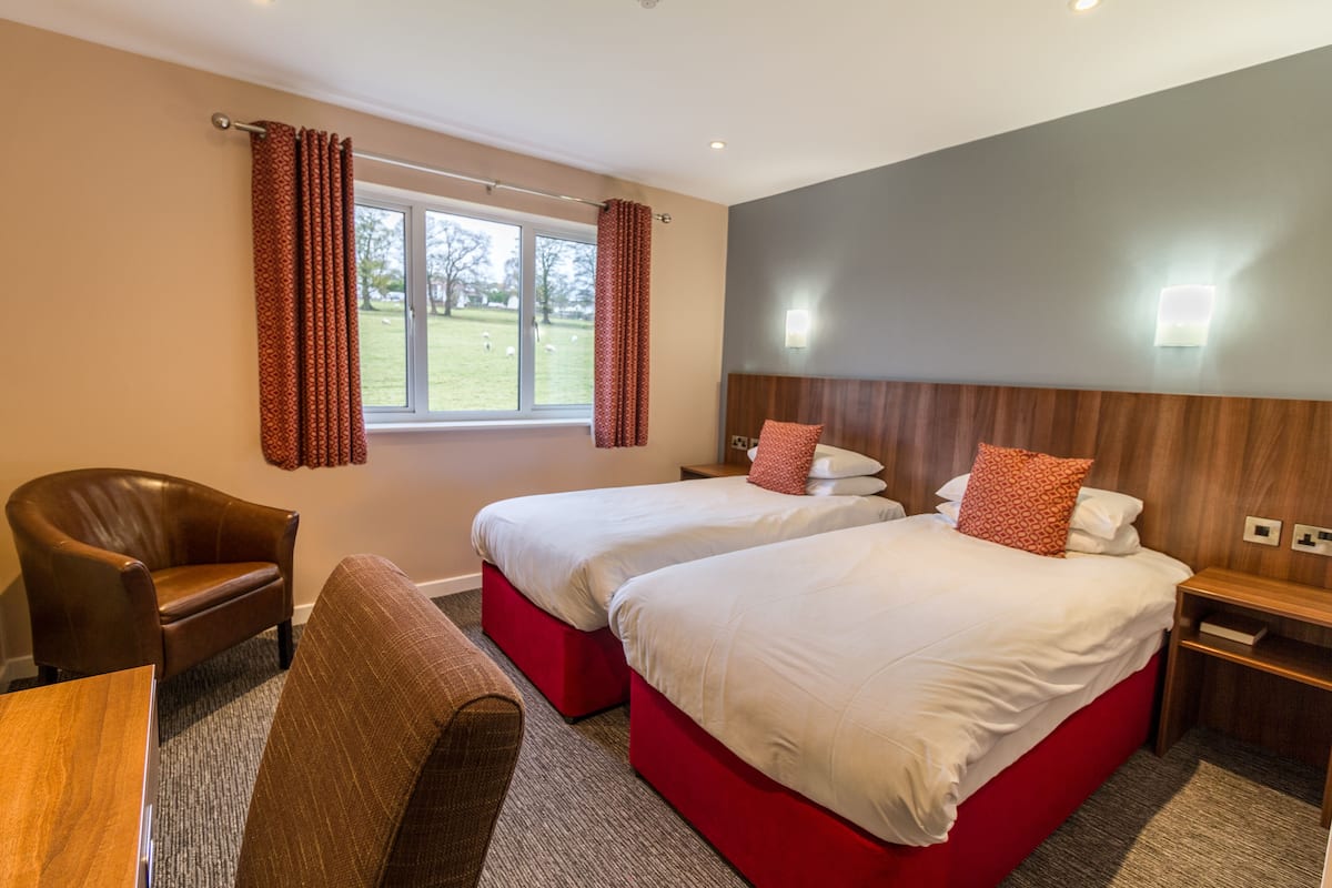 Habitación de hotel con dos camas, una mesita de noche de madera, un sillón de cuero y vista al exterior a través de una ventana.