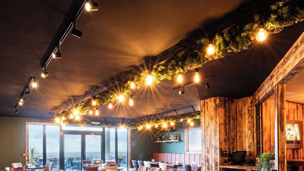 A restaurant with wooden flooring, multiple tables and chairs, string lights, and large windows offering a view.