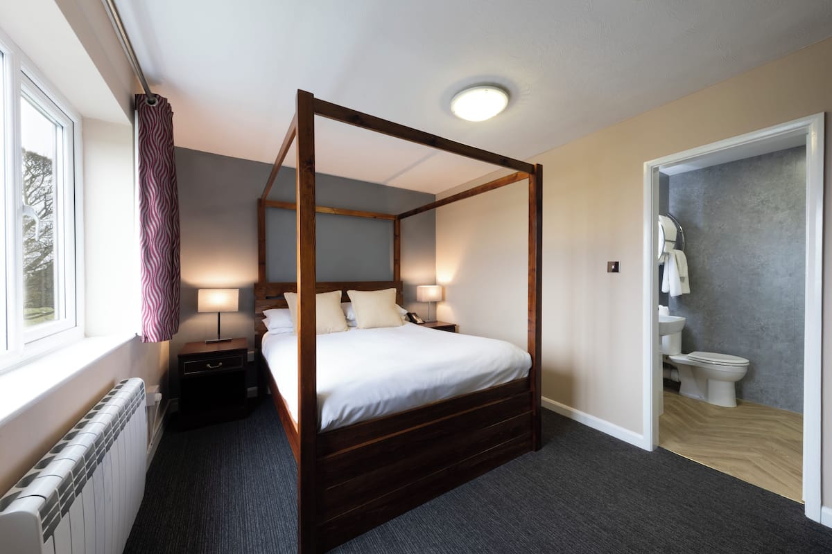 Una cama con dosel de madera, dos mesitas de noche, una ventana con cortina, un baño con inodoro y lavabo, y un radiador.