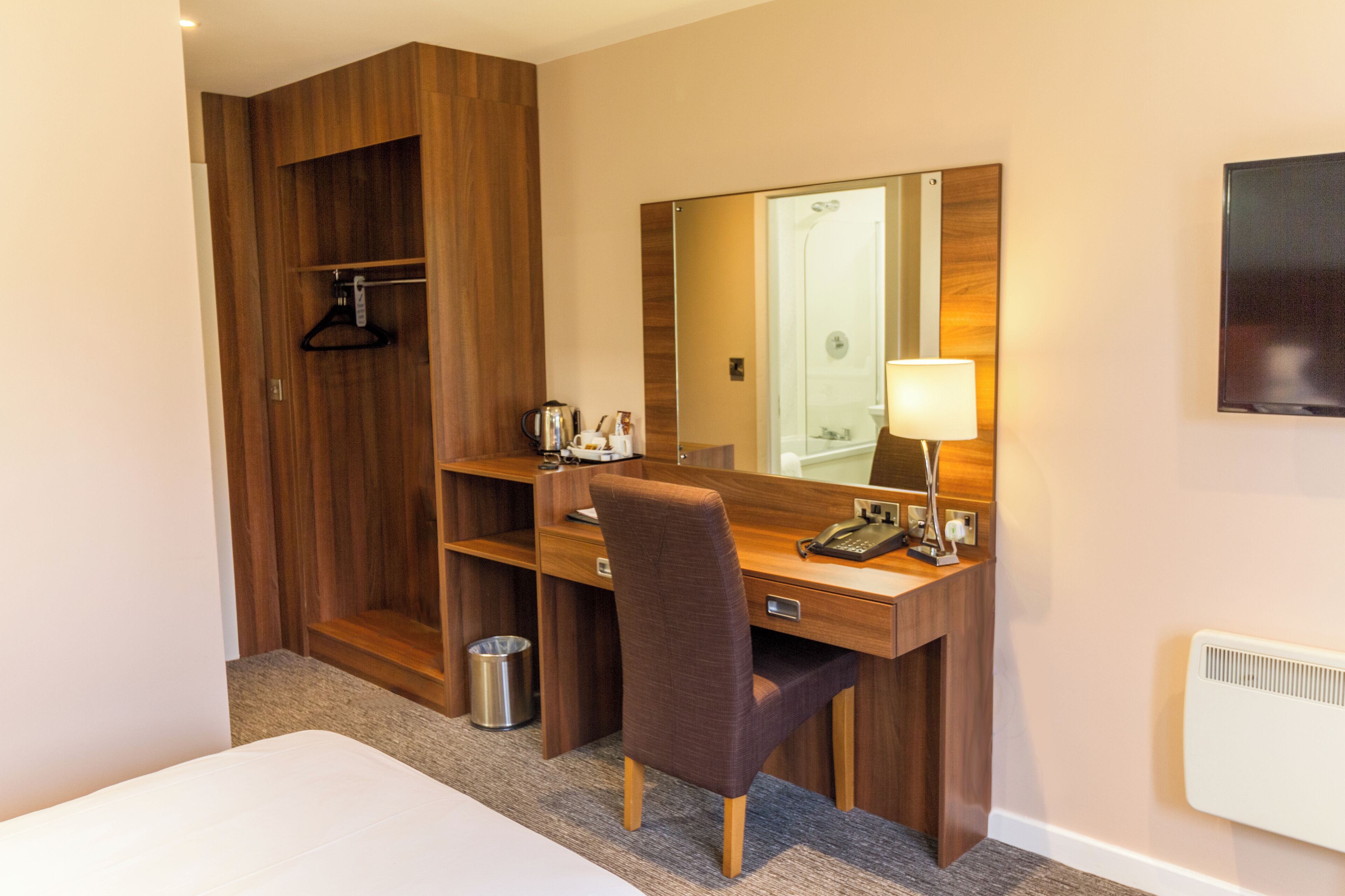 Une chambre d’hôtel avec un bureau en bois, une chaise, un miroir, une lampe, un téléphone et un radiateur.