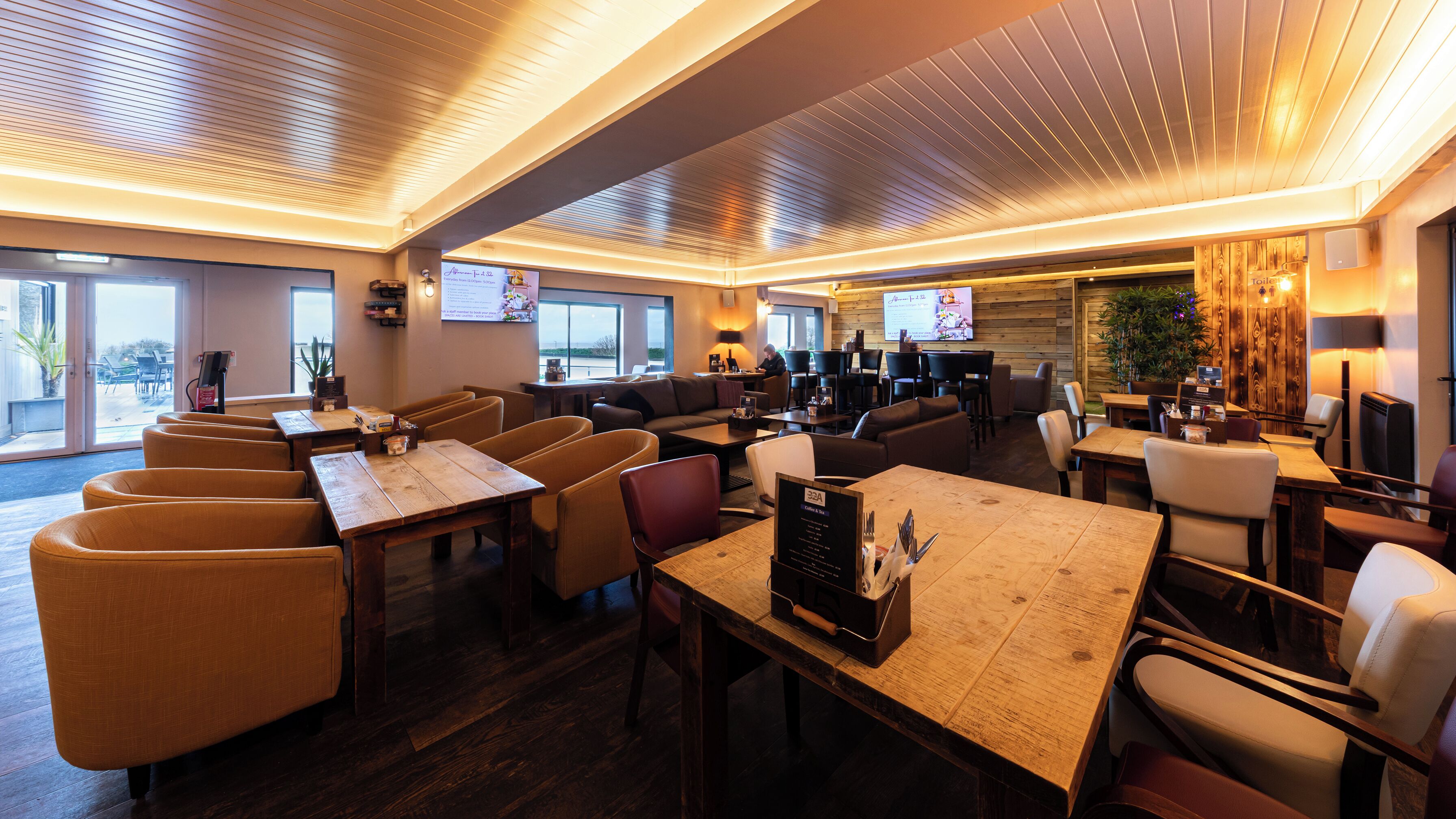 A spacious hotel lobby with multiple seating areas, including sofas and tables, and a large television screen displaying a menu.