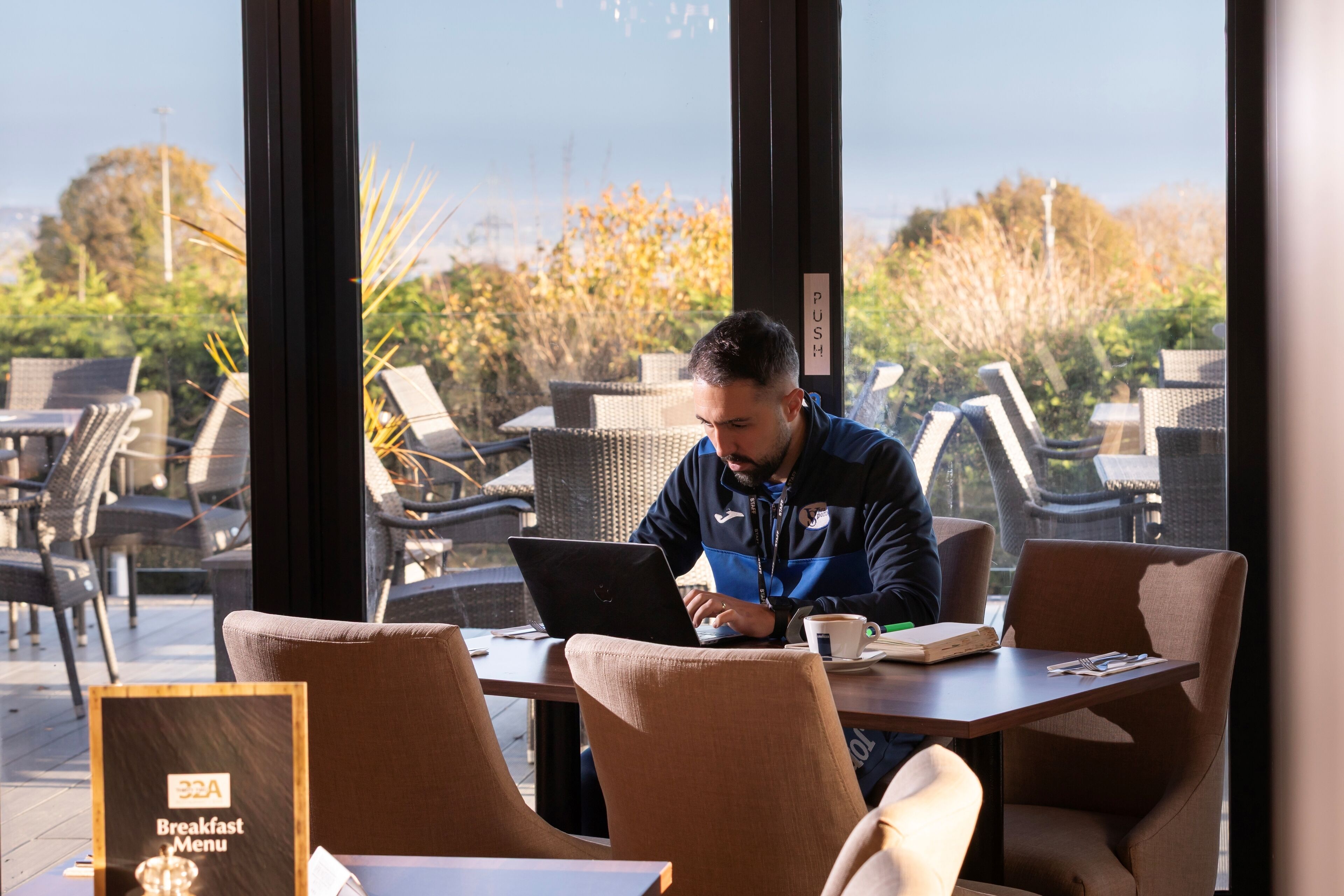 A man working on a laptop at a table with a view of outdoor seating and greenery.