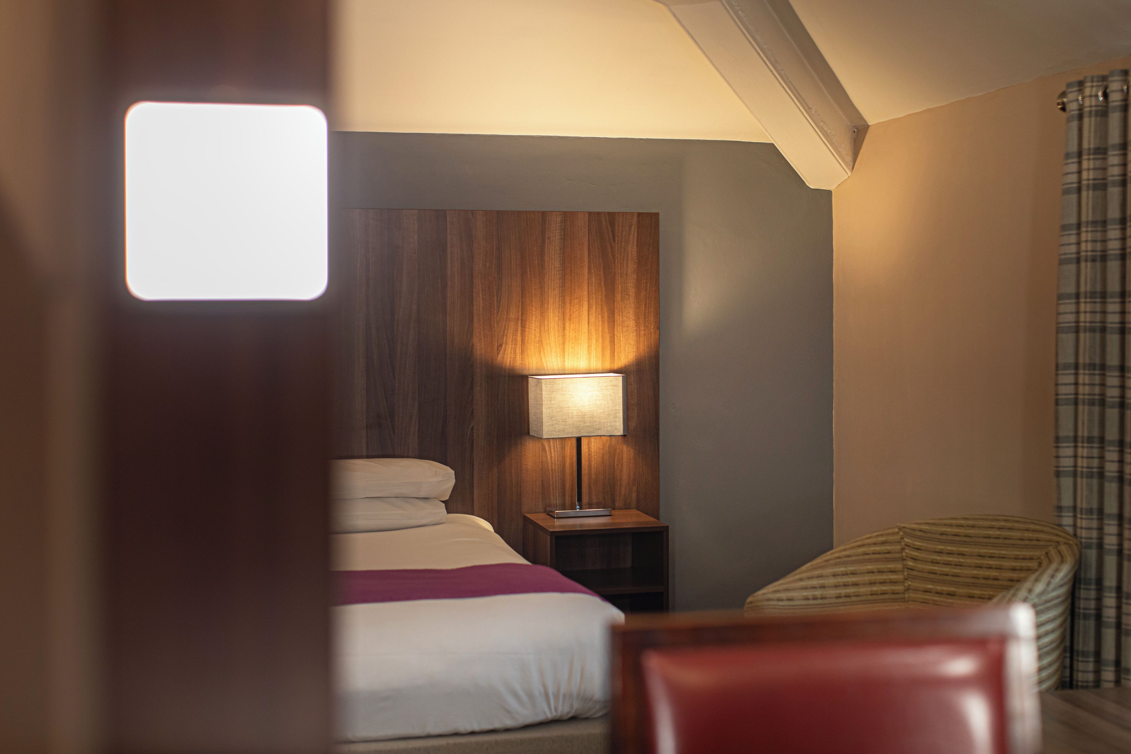 Quarto de hotel com cama, mesa de cabeceira com abajur, cadeira e janela com cortinas.