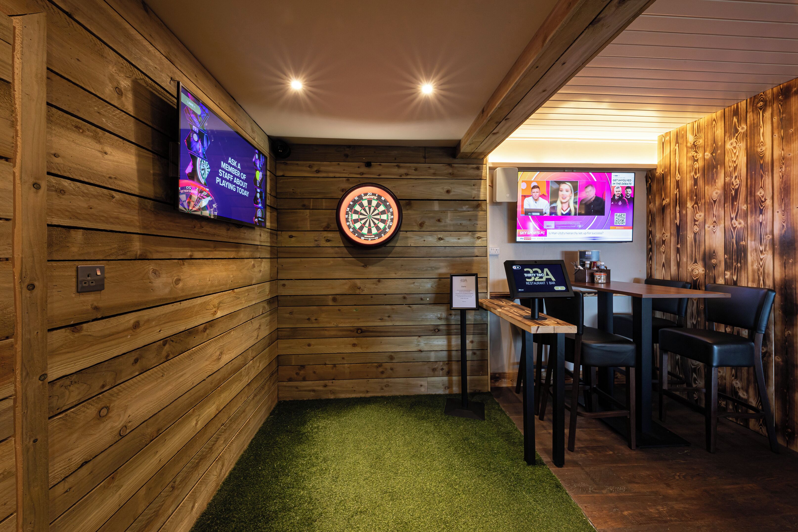 A room with wooden walls, a dartboard, a TV screen, and a table with chairs.