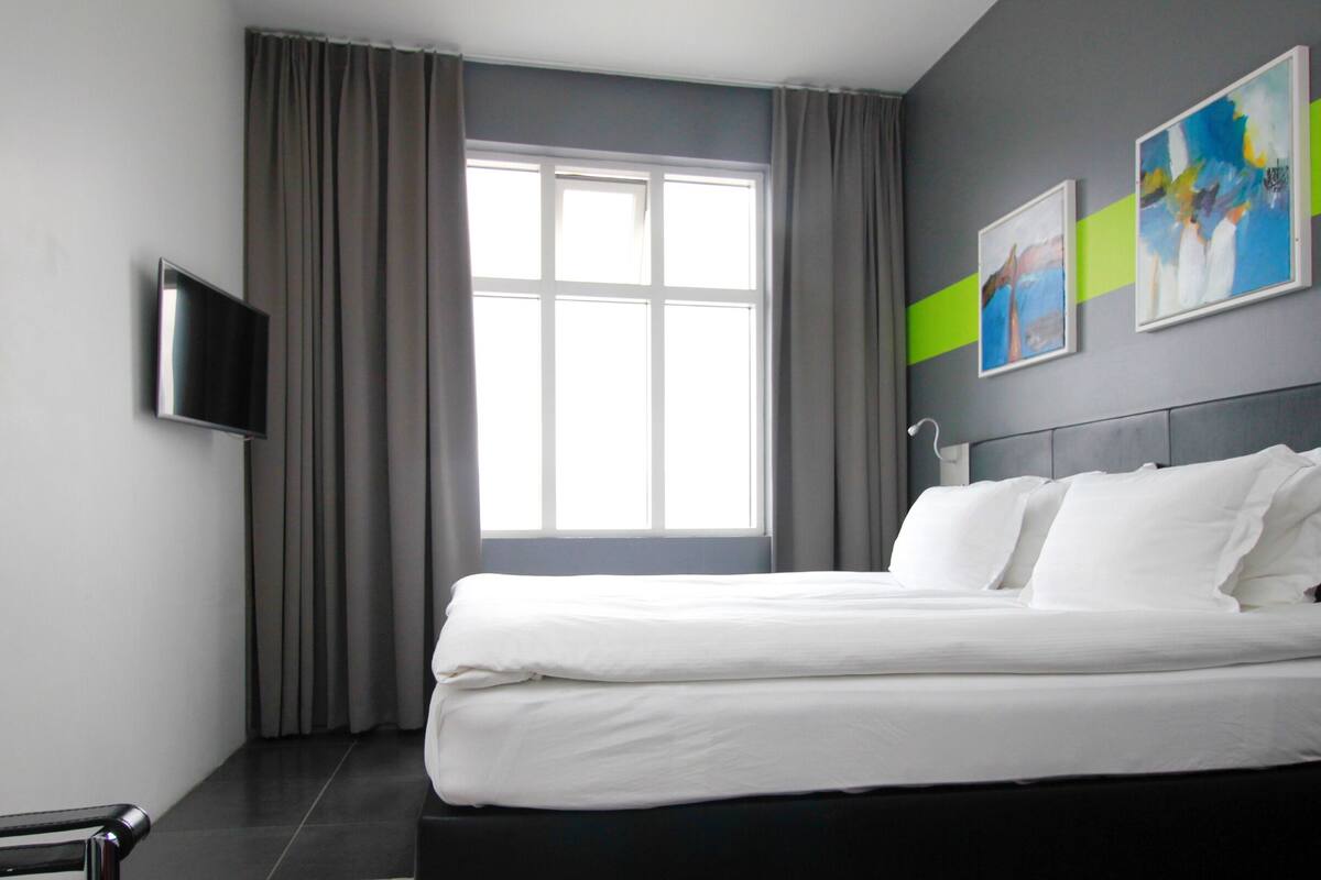 Een moderne hotelkamer met een groot bed, grijze gordijnen, een flatscreen televisie aan de muur en twee abstracte schilderijen.