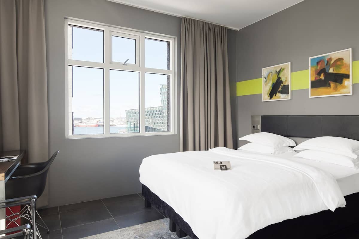 Een hotelkamer met een bed, een bureau en een stoel. Er is een raam met gordijnen, en twee schilderijen aan de muur.
