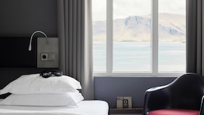 Habitación de hotel con cama, una silla, vistas a un lago y montañas, y un termostato en la pared.
