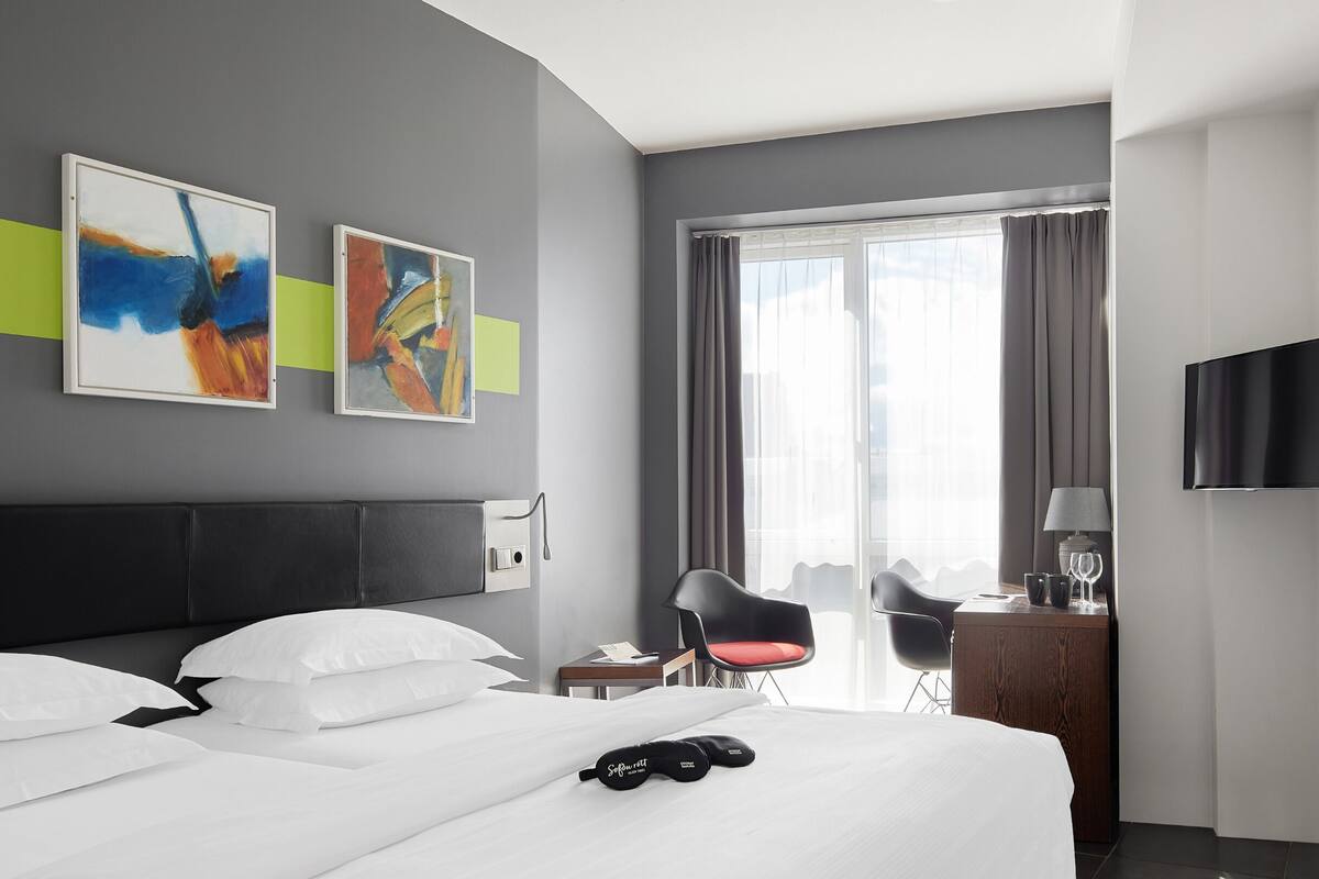 Een moderne hotelkamer met een groot bed, twee schilderijen aan de muur, een bureau met lamp, een stoel en een televisie.